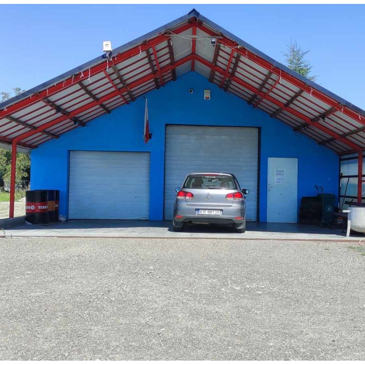 Szary samochód osobowy zaparkowany przed niebieskim budynkiem warsztatu z dwoma bramami garażowymi i czerwonym dachem, widoczne beczki Texaco i flaga Polski.