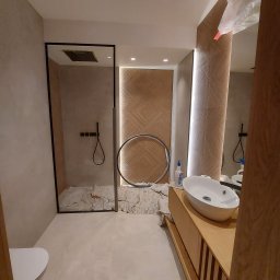 Matbau - Nowoczesna łazienka w trakcie wykończenia: widoczna kabina prysznicowa typu walk-in z czarną ramą, dekoracyjna ściana z drewnianą mozaiką, okrągłe lustro oprawione w czarną ramę oraz umywalka...