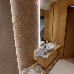 Matbau - Nowoczesna łazienka w trakcie remontu: drewniane panele ścienne z geometrycznym wzorem, okrągła umywalka na drewnianej szafce, widoczne narzędzia i środki czystości, odzwierciedlenie osoby w lustrze.