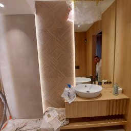 Matbau - Nowoczesna łazienka w trakcie wykończenia, z drewnianą okładziną ścienną w geometryczny wzór, okrągłą umywalką nablatową i widocznym pracownikiem w lustrze.