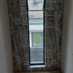 Matbau - Montaż płytek imitujących marmur wokół okna na klatce schodowej, widoczne krzyżyki dystansowe i niezabezpieczone kable.
