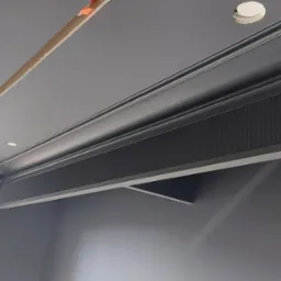 Szare wnętrze w trakcie remontu, widoczny sufit z listwami dekoracyjnymi i otworem na oświetlenie oraz fragment okna.