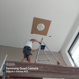 Matbau - Mężczyzna na drabinie przygotowuje sufit do montażu oświetlenia, widoczna płyta kartonowo-gipsowa z wyciętym otworem i wystającymi przewodami.