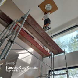 Matbau - Pracownik na drabinie, montuje oświetlenie w suficie podwieszanym, widoczne rusztowanie i duże okno z widokiem na drzewa.