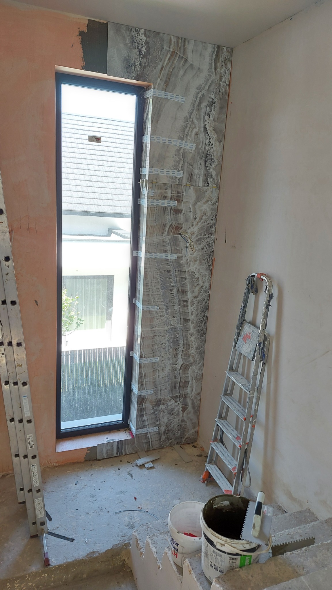 Montaż płytek imitujących marmur wokół okna, widoczne listwy poziomujące, drabiny i wiaderka z zaprawą na podłodze w trakcie prac wykończeniowych.