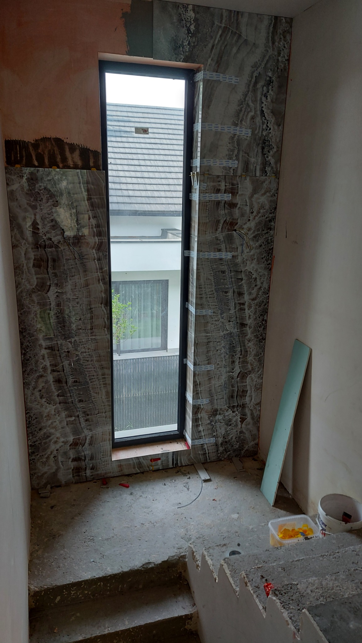 Montaż płytek imitujących marmur wokół okna na klatce schodowej, widoczne taśmy montażowe, surowy stan ścian i schodów, w tle widok na dom przez okno.