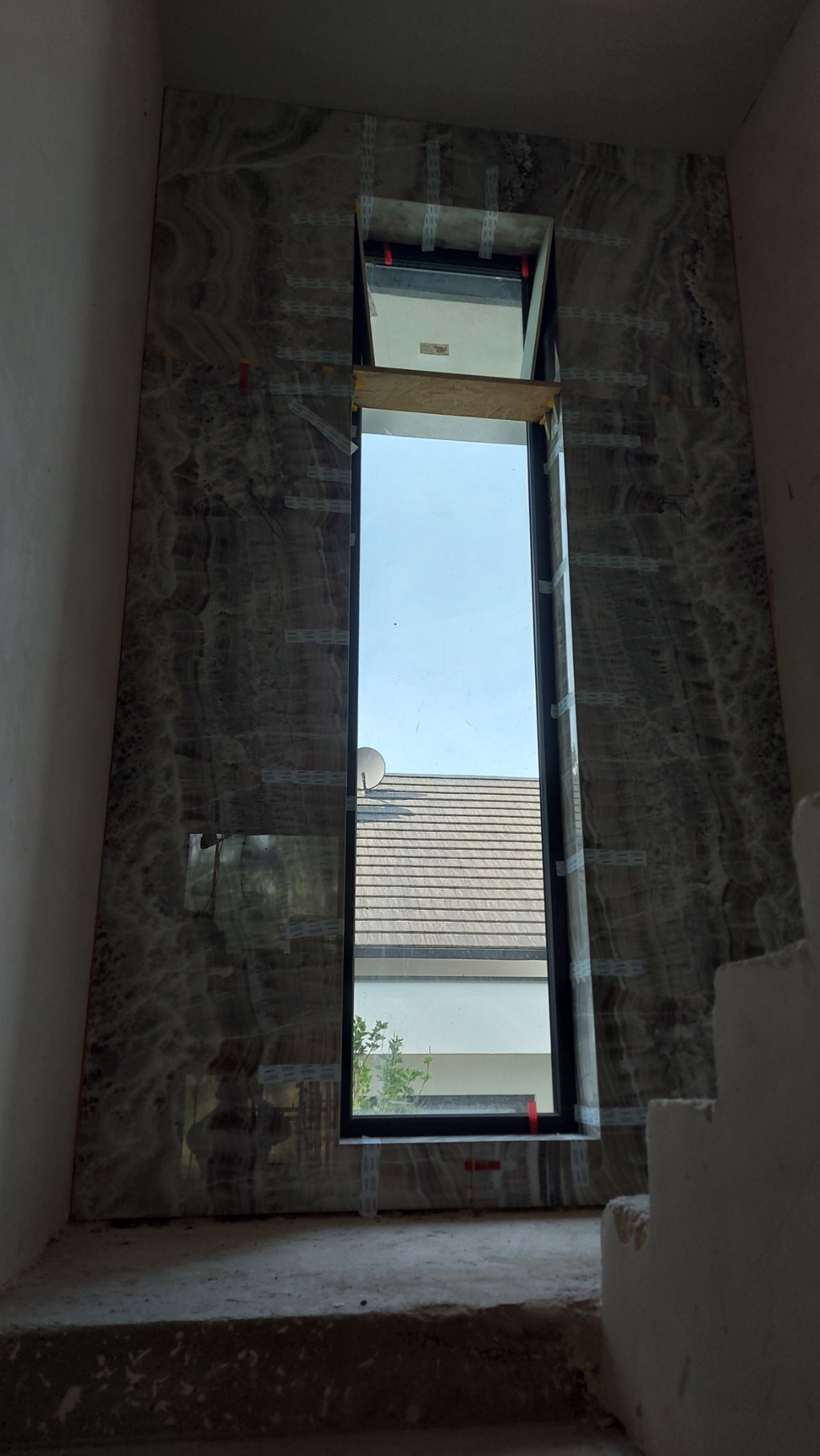 Montaż płytek imitujących marmur wokół okna w trakcie remontu, widoczne taśmy mocujące i fragment schodów.