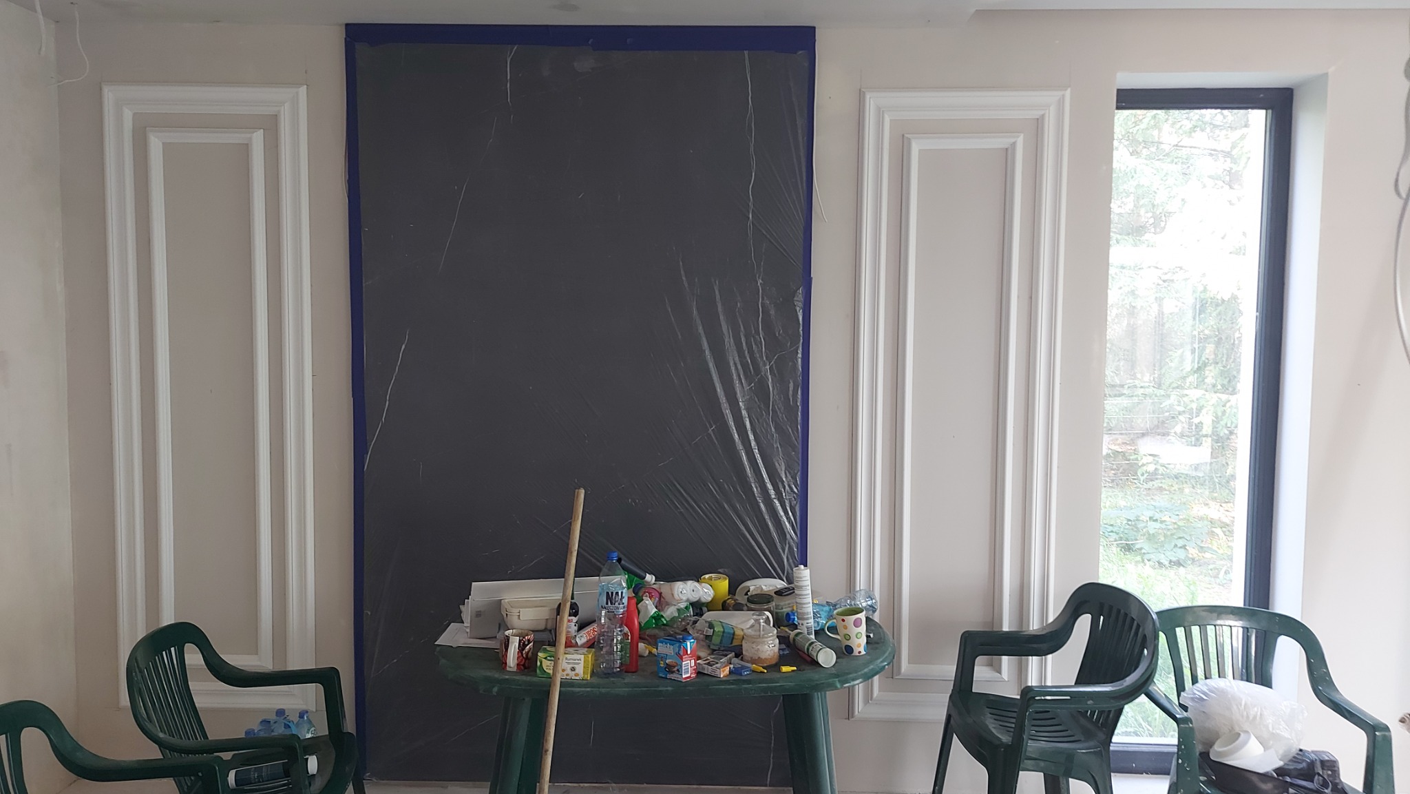 Przygotowanie do remontu: ściana zabezpieczona folią malarską, dekoracyjne listwy ścienne, stół z narzędziami i materiałami, krzesła ogrodowe.