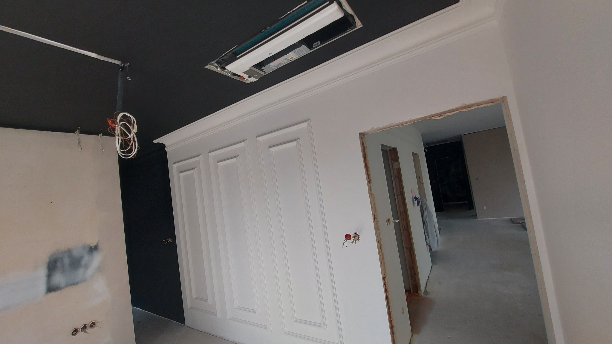 Wnętrze w trakcie remontu: białe panele ścienne z dekoracyjnymi frezami, czarny sufit z otwartą instalacją oświetleniową, widoczne przewody elektryczne i surowe wykończenie ościeżnicy drzwiowej.