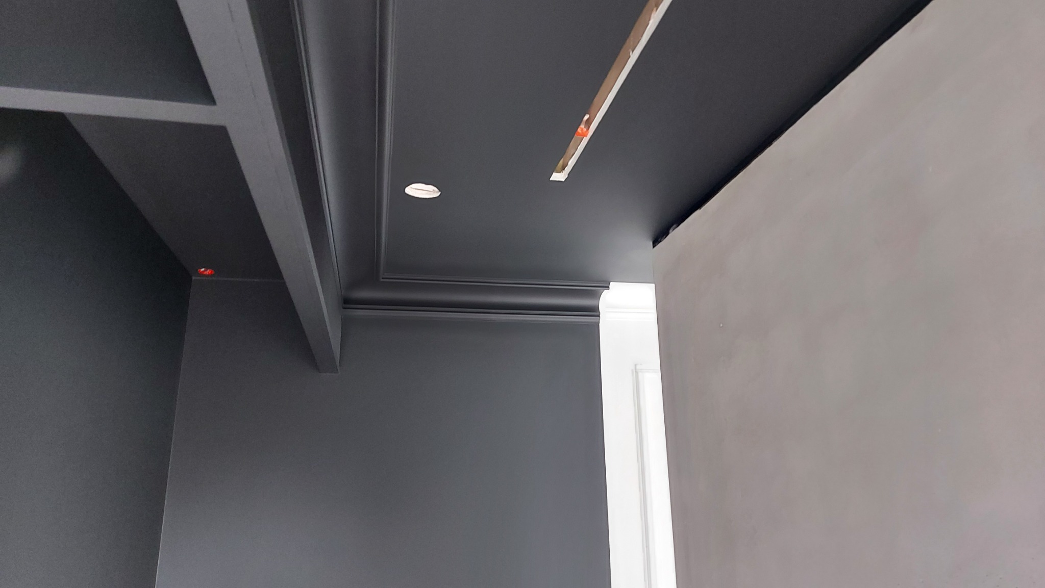 Ciemnoszara ściana i sufit z ozdobnymi listwami, widoczne ślady po szpachlowaniu na sąsiedniej ścianie, zamontowane oświetlenie punktowe w suficie.