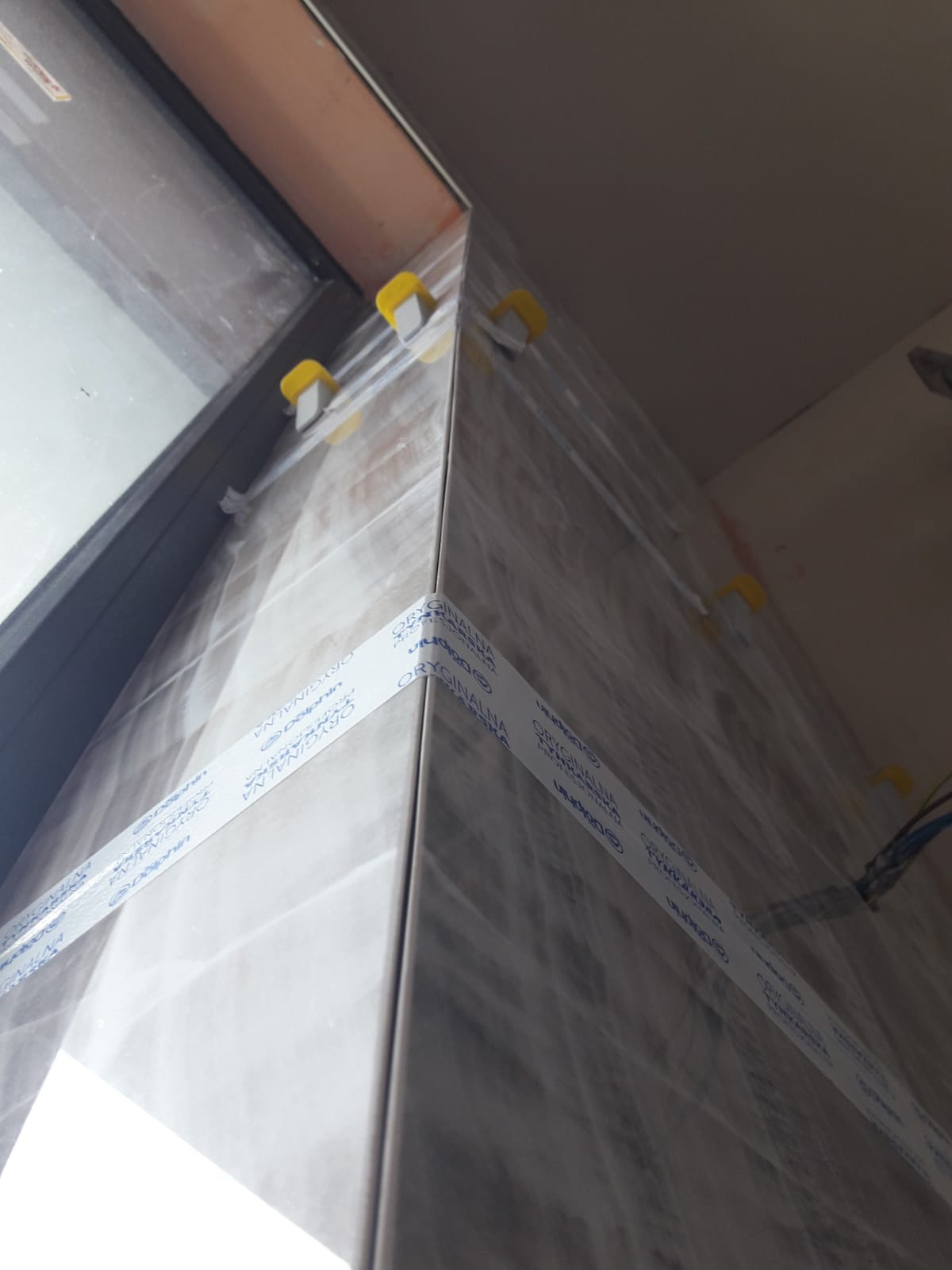 Świeżo ułożone, szare płytki ceramiczne na ścianie, zabezpieczone taśmą z napisem 'ORYGINALNA RECEPTURA POLSKA' i poziomowane żółtymi klipsami, widok z dołu w rogu pomieszczenia, z widocznym...