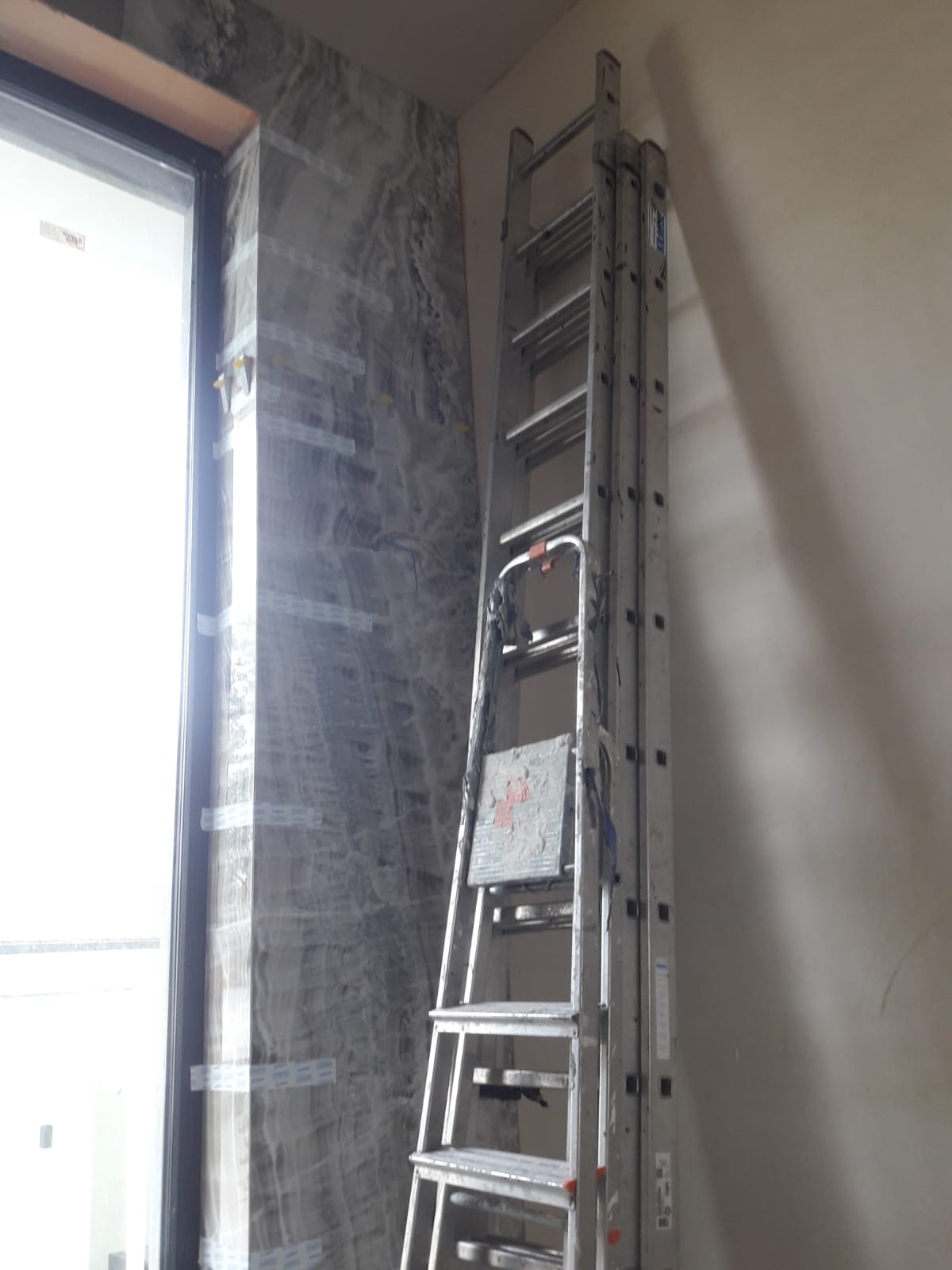 Ściana wykończona płytami imitującymi marmur, zabezpieczona taśmą, z ustawioną aluminiową drabiną. Widok na okno.