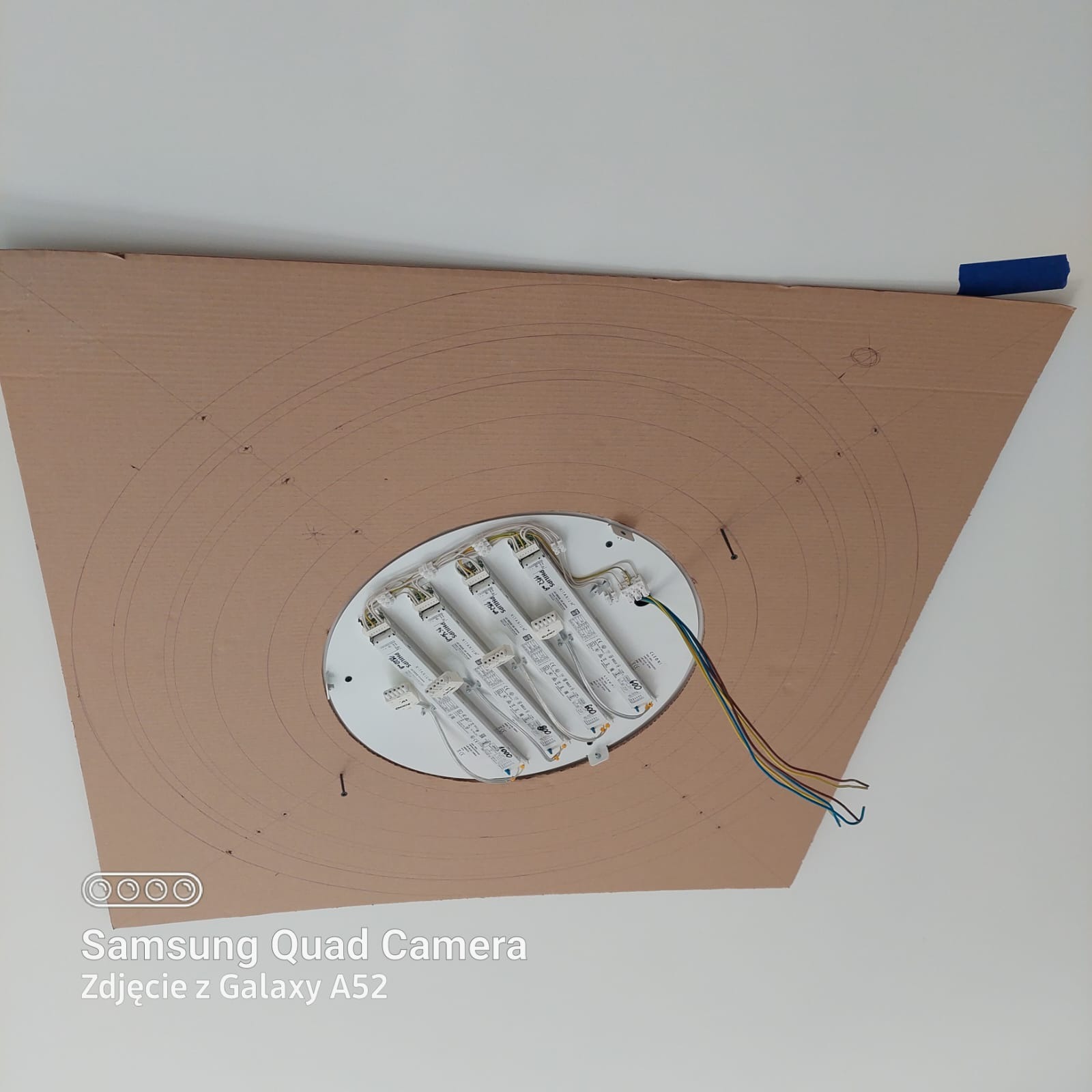 Przygotowanie do montażu oświetlenia sufitowego: widoczna płyta kartonowa z odrysowanymi okręgami i wyciętym otworem na lampę, z zamontowanym modułem LED Philips i wystającymi kablami, przyklejona...