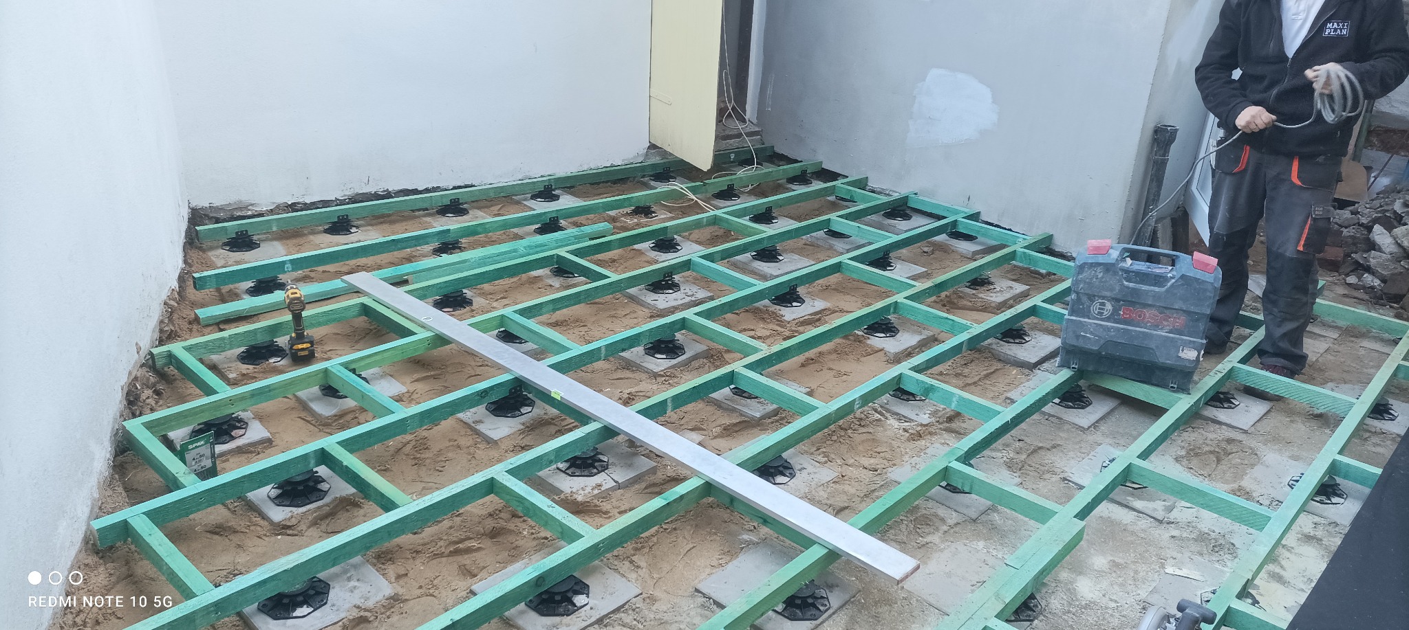 Konstrukcja szkieletowa pod taras z regulowanymi wspornikami, widoczna poziomica i skrzynka narzędziowa Bosch, w tle ściana budynku.