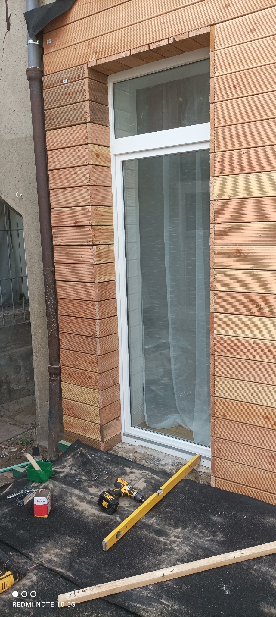 Montaż drewnianej elewacji wokół okna, widoczne deski elewacyjne, poziomica, wkrętarka i narzędzia na czarnej macie zabezpieczającej.