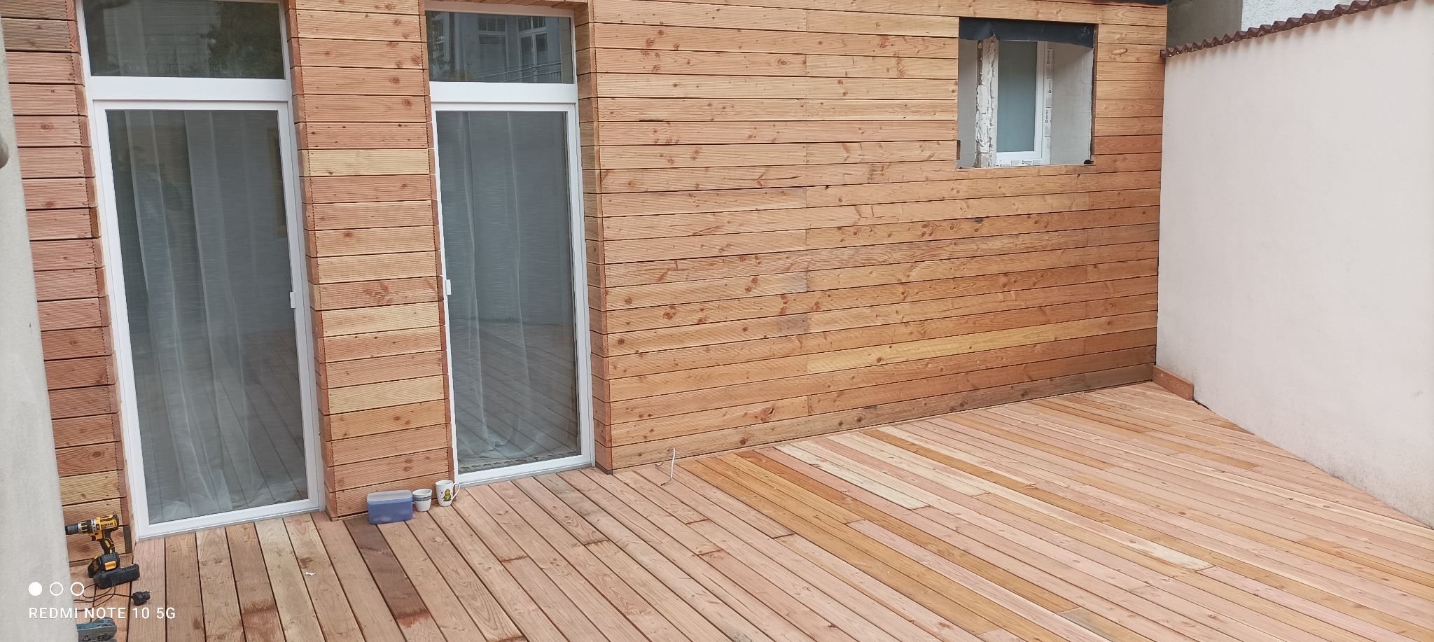 Wykończony taras z desek drewnianych, widoczne dwie białe ramy okienne i fragment ściany z oknem w trakcie montażu, w rogu wiertarka akumulatorowa i pojemnik z narzędziami.