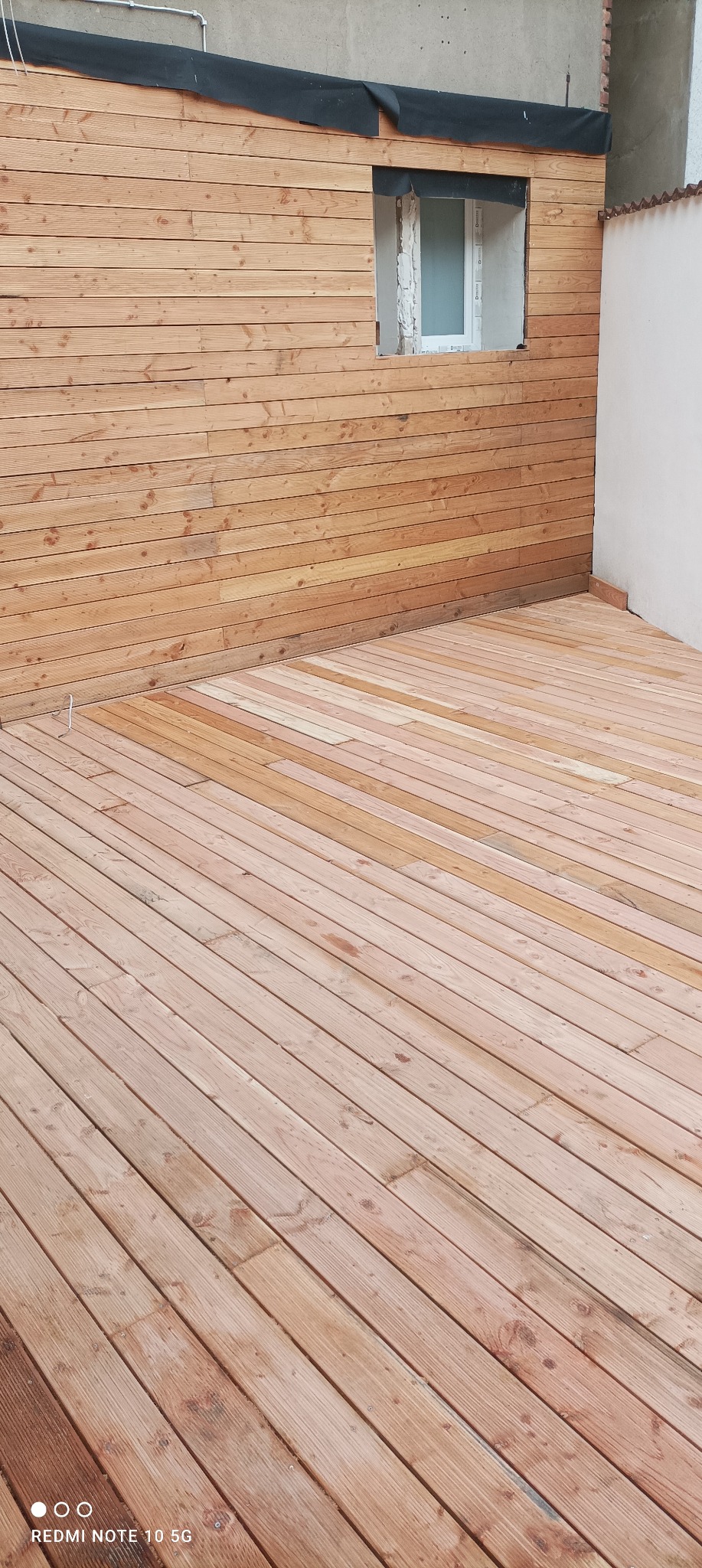 Drewniany taras z deskami ułożonymi pod kątem, przylegający do ściany z drewnianą boazerią i oknem w stanie surowym.