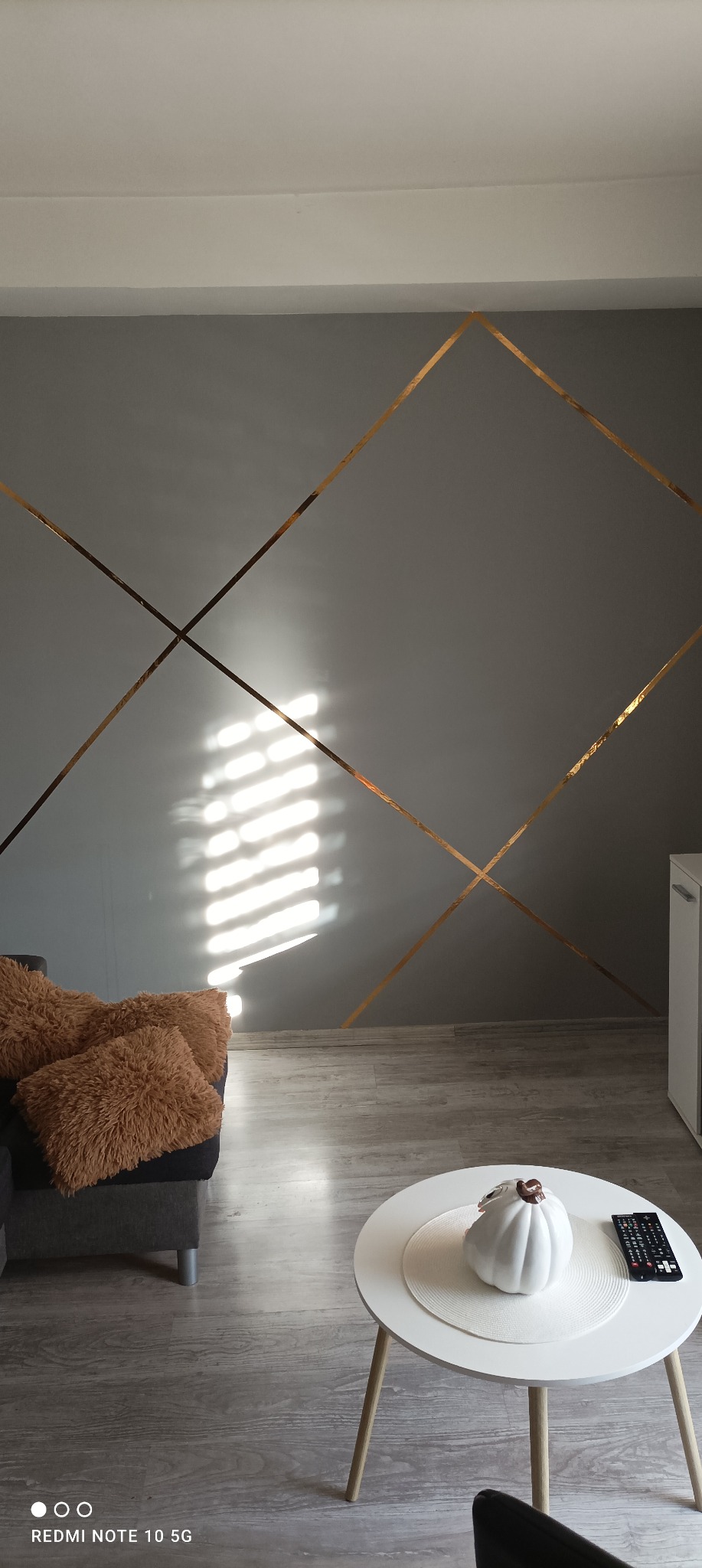 Szara ściana w salonie z dekoracyjnymi, złotymi paskami tworzącymi geometryczny wzór, oświetlona promieniami słonecznymi, widoczny fragment szarej sofy z brązowymi poduszkami i okrągły biały stolik...