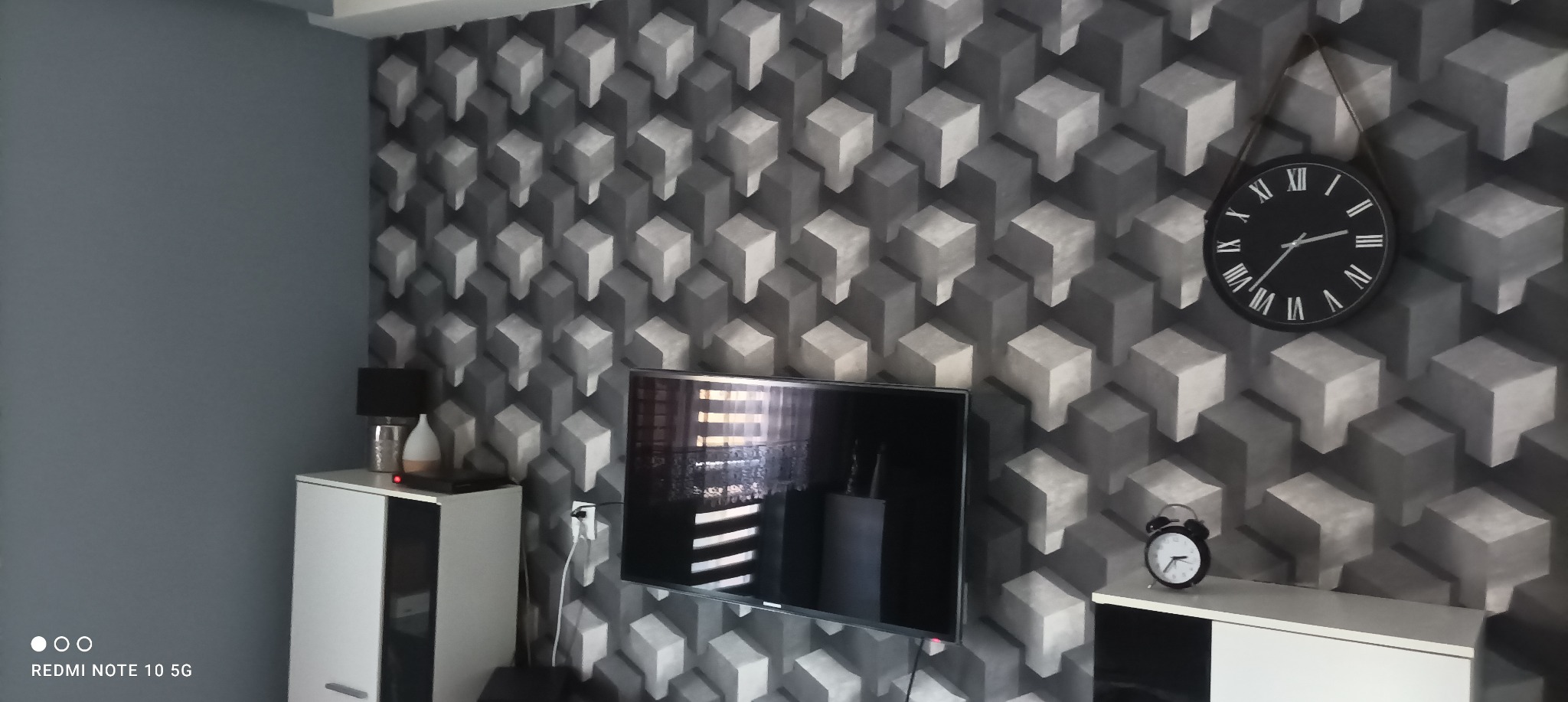Pokój z szarą ścianą i tapetą w geometryczne wzory 3D, na ścianie wiszący zegar i telewizor, na szafkach lampka nocna i zegarek.