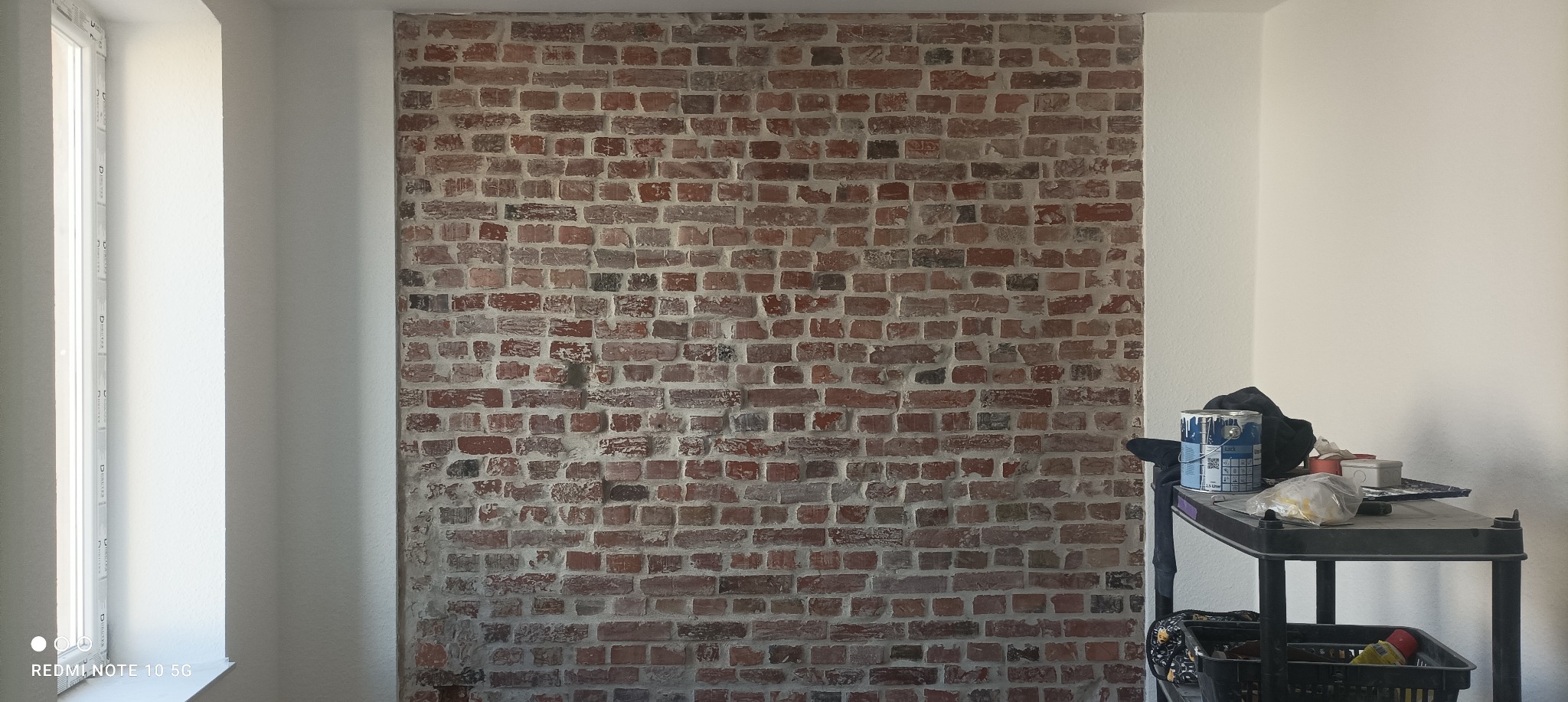 Fragment ściany z dekoracyjnej cegły w trakcie prac wykończeniowych, widoczny wózek z puszkami farby i narzędziami malarskimi, ściany pomalowane na biało, okno po lewej stronie.