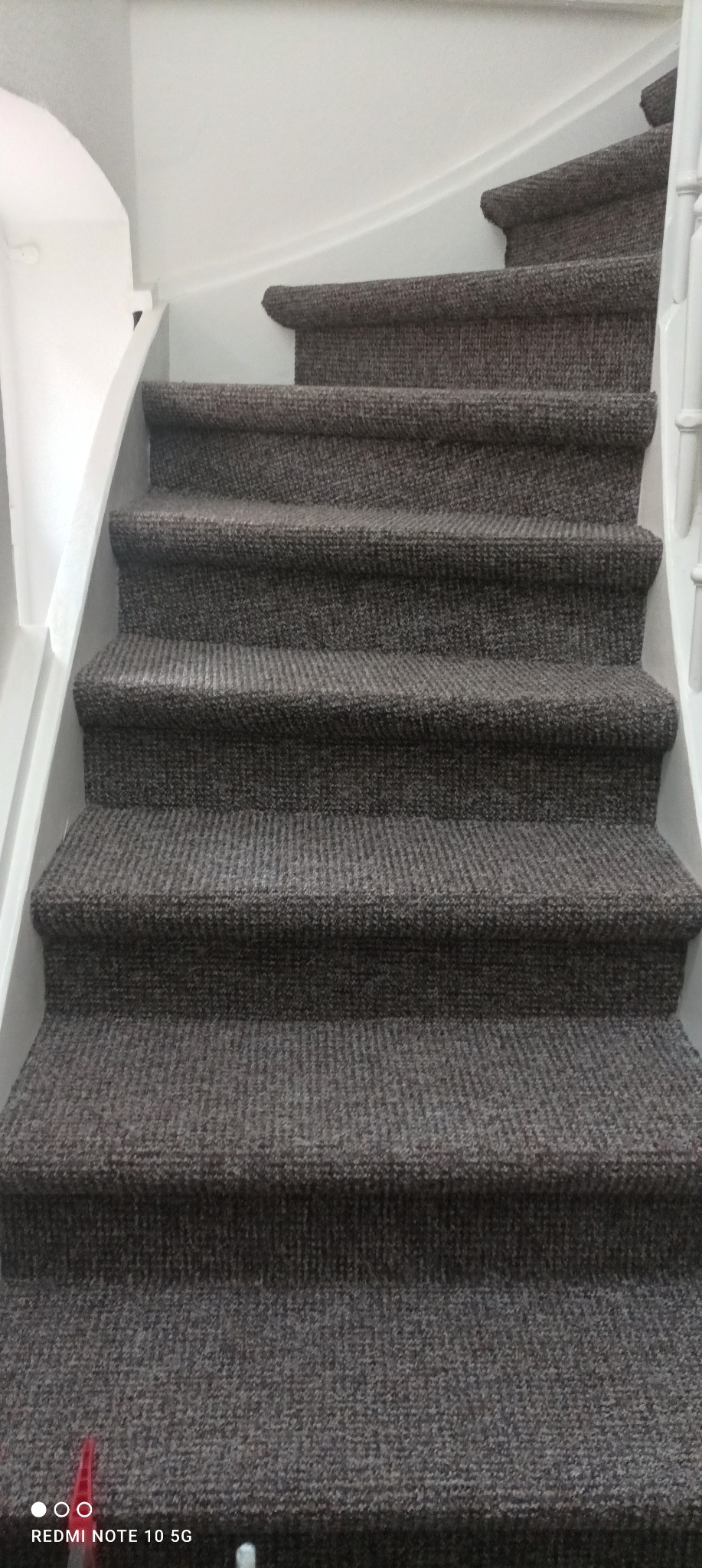 Schody wewnątrz budynku, wyłożone szarym dywanem, z białymi barierkami po bokach.