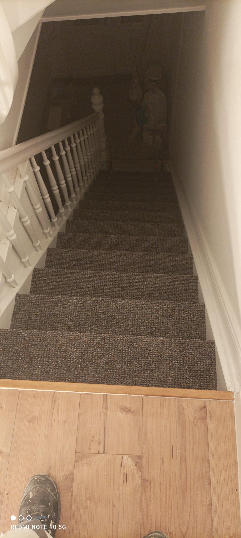Widok z góry na schody wyłożone brązową wykładziną dywanową, z drewnianą podłogą na dole i białymi balustradami po bokach, w tle widoczny korytarz w słabym oświetleniu.