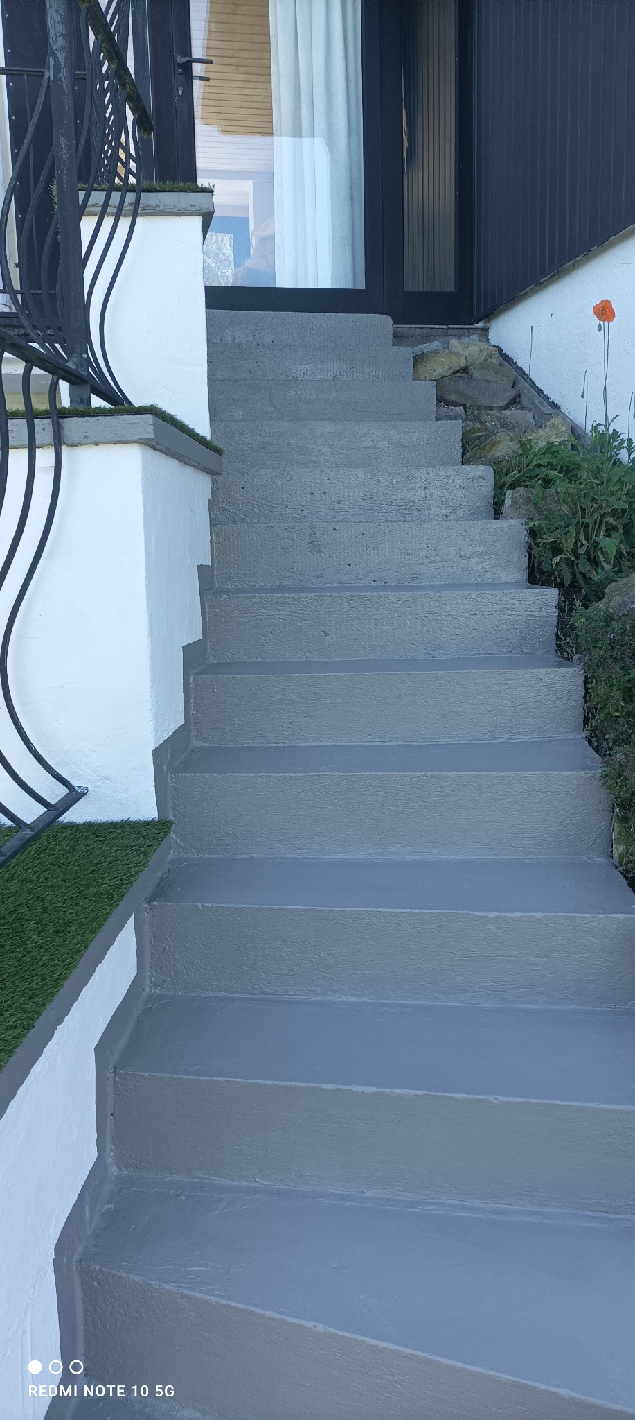 Betonowe schody zewnętrzne pomalowane na szaro, z fragmentem białej ściany, czarną balustradą i sztuczną trawą na krawędzi.