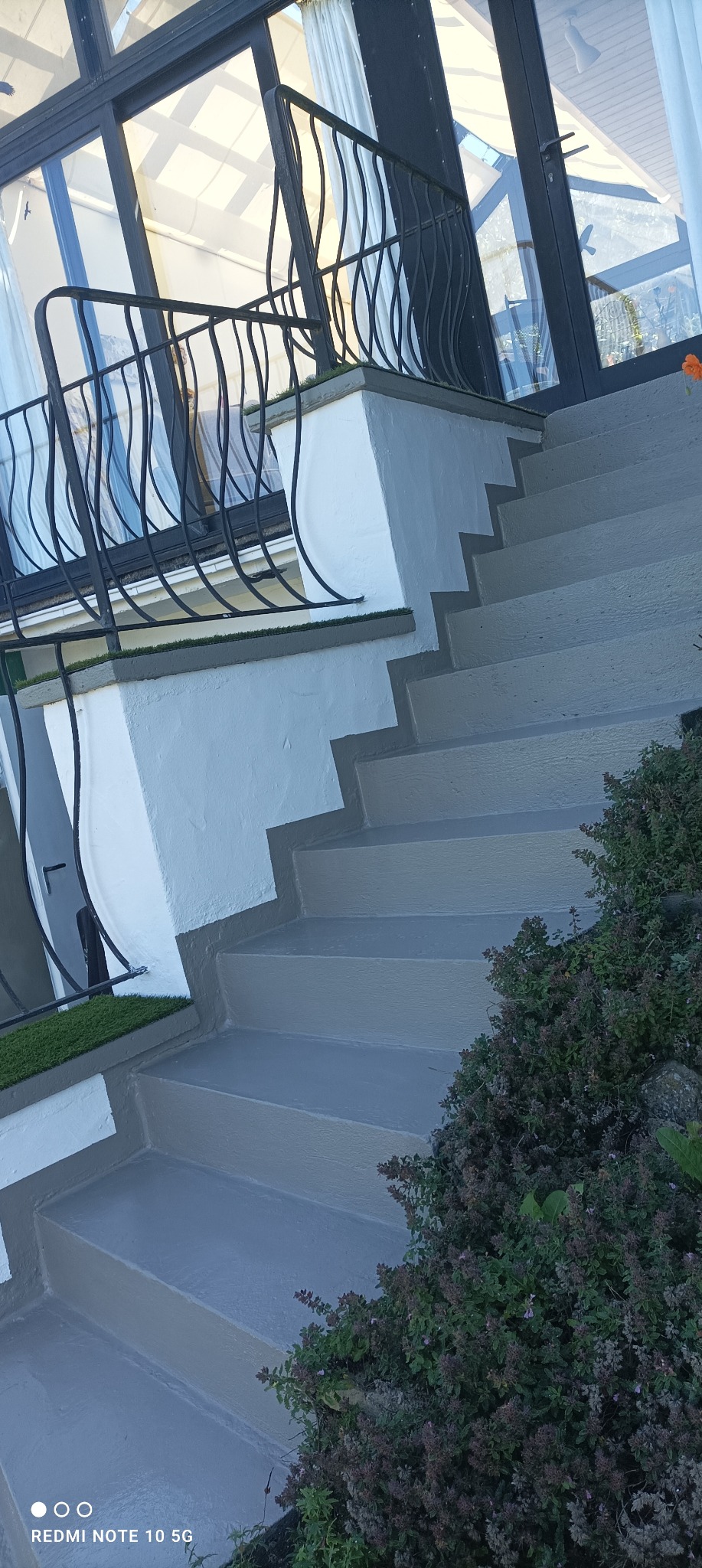Zewnętrzne schody prowadzące do przeszklonego budynku, świeżo pomalowane na szaro, z ozdobną, czarną balustradą i fragmentami sztucznej trawy.