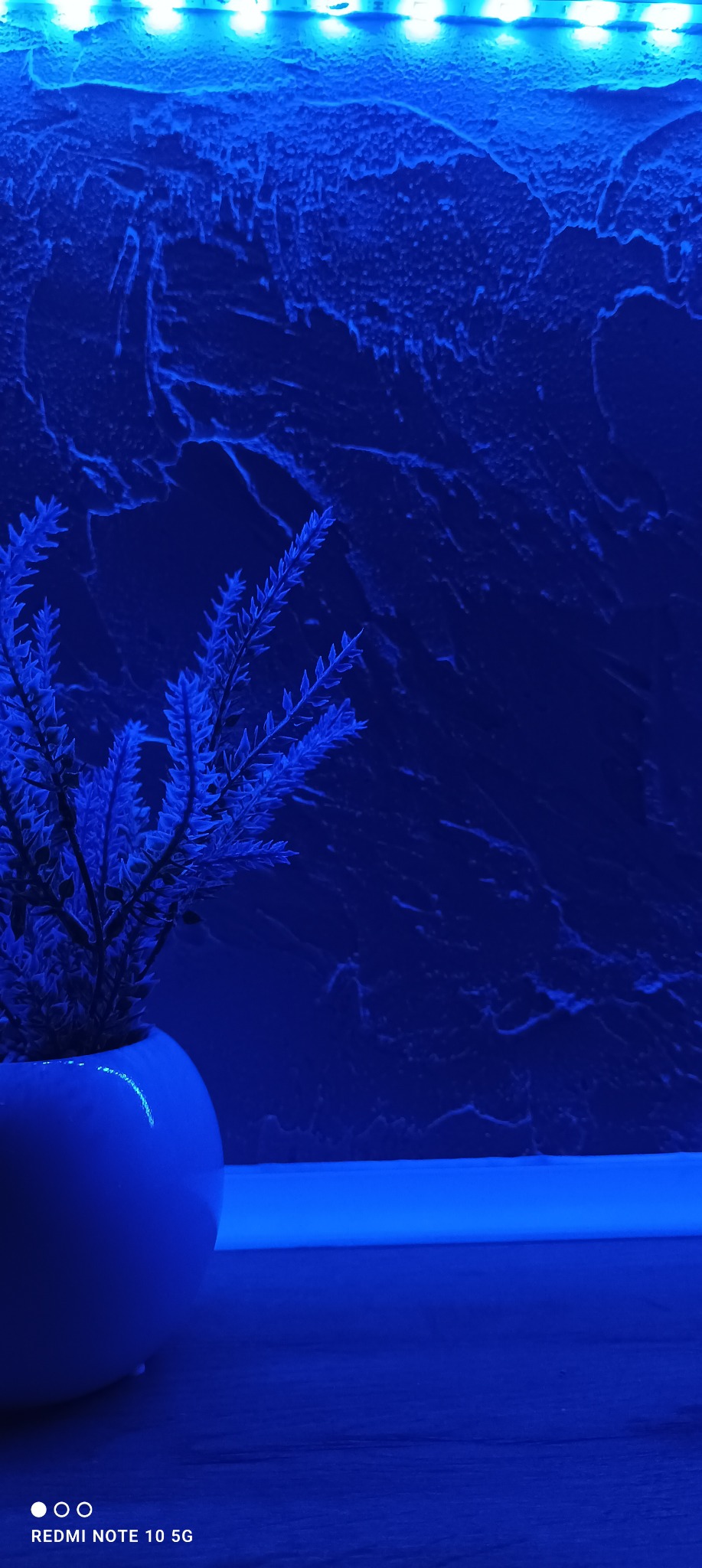 Niebieska ściana z dekoracyjną strukturą, oświetlona listwą LED, z rośliną w białej doniczce, tworząca nowoczesny wystrój wnętrza.