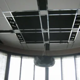 Sufit podwieszany z czarnymi panelami akustycznymi, oświetleniem punktowym i metalowymi listwami dekoracyjnymi, widok z dołu na okrągły pokój z żaluzjami i lampą wiszącą.