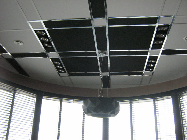 Sufit podwieszany z czarnymi panelami akustycznymi, oświetleniem punktowym i metalowymi listwami dekoracyjnymi, widok z dołu na okrągły pokój z żaluzjami i lampą wiszącą.