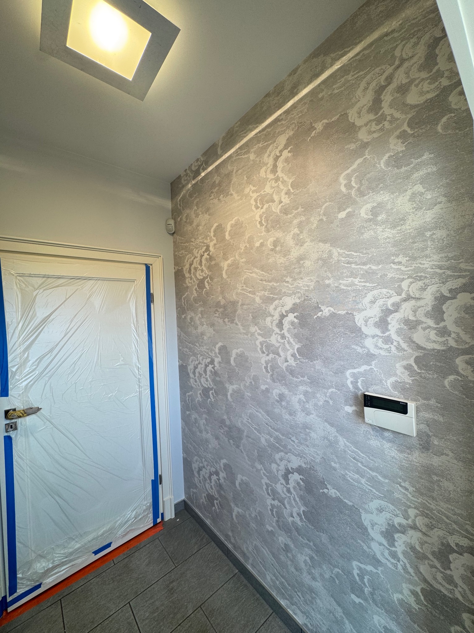 Ściana z wzorzystą tapetą w odcieniach szarości, zabezpieczone drzwi z futryną. Widoczny fragment podłogi z szarych płytek i panel sterowania na ścianie.