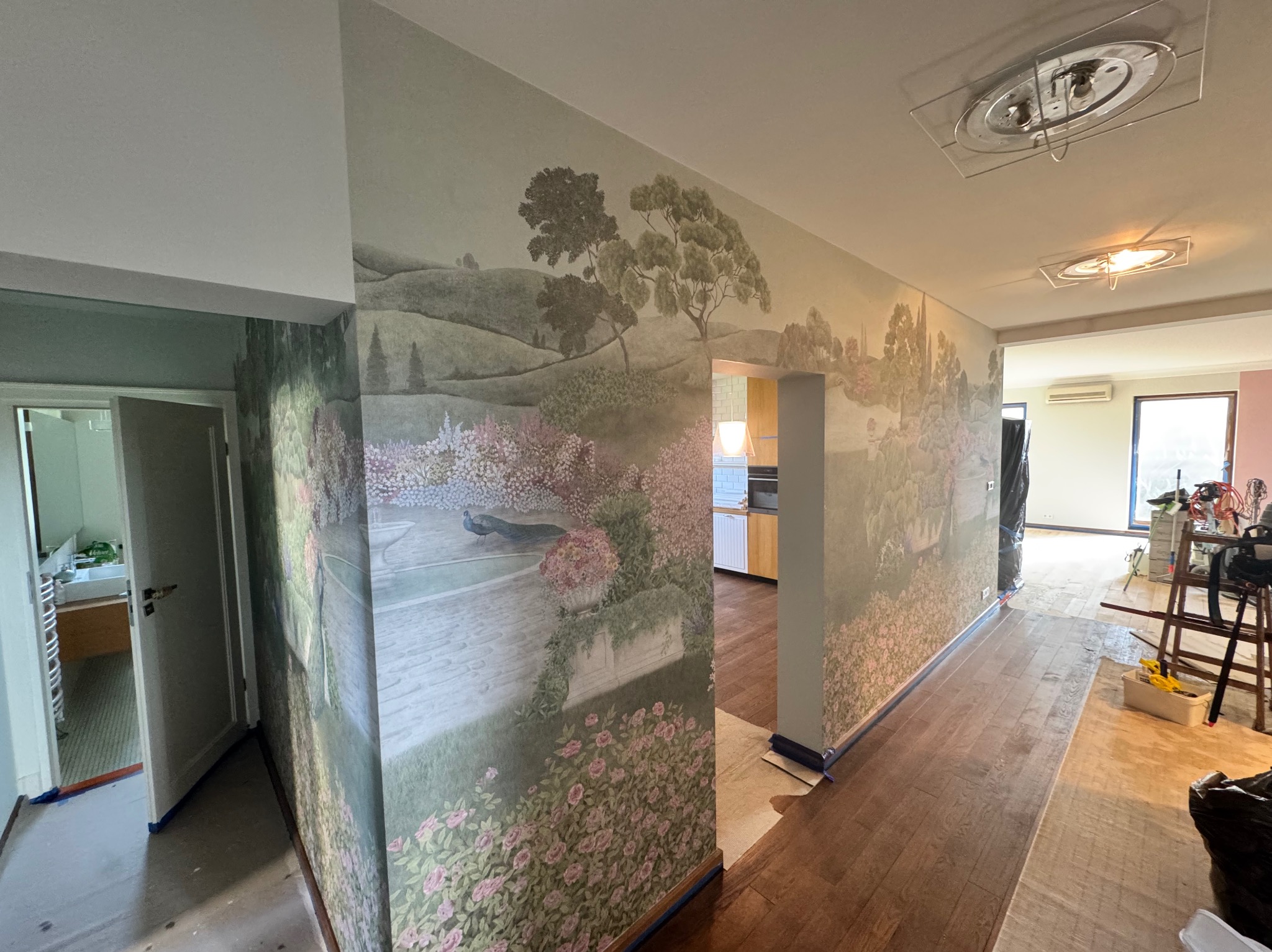 Wnętrze po dekoracji ścian fototapetą z motywem parku i pawia. Widoczne fragmenty pomieszczeń, zabezpieczona podłoga, oświetlenie sufitowe.