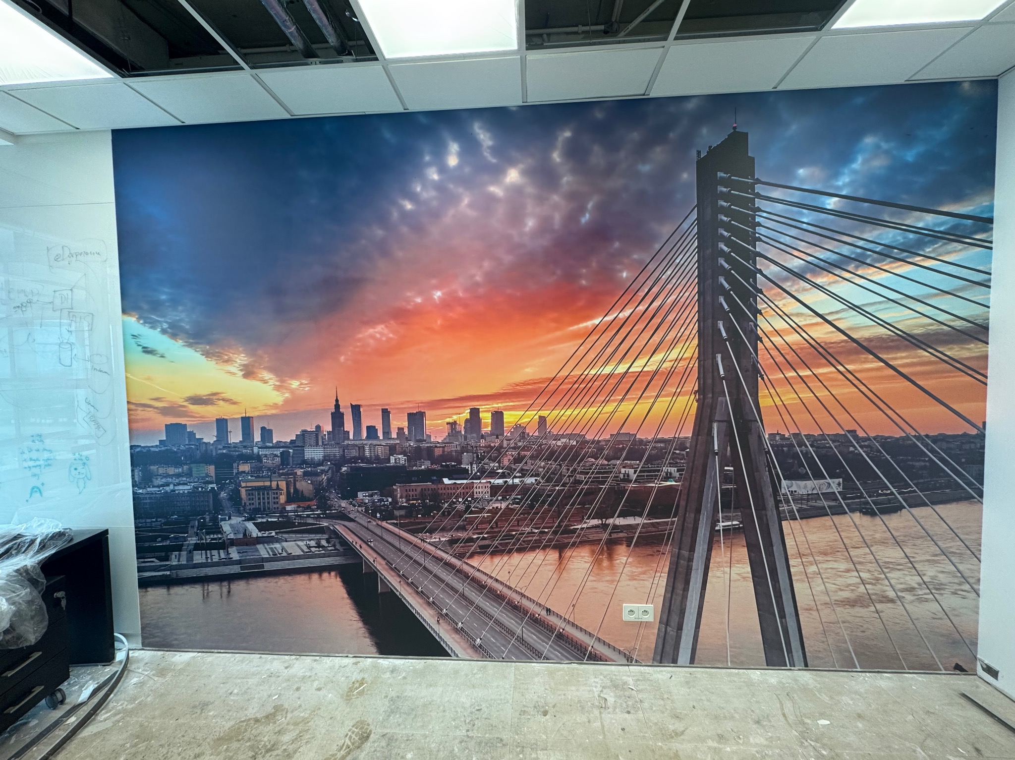 Fototapeta z widokiem na Warszawę i most Świętokrzyski, naklejona na ścianie biura. Widoczne gniazdka elektryczne i fragment sufitu podwieszanego.