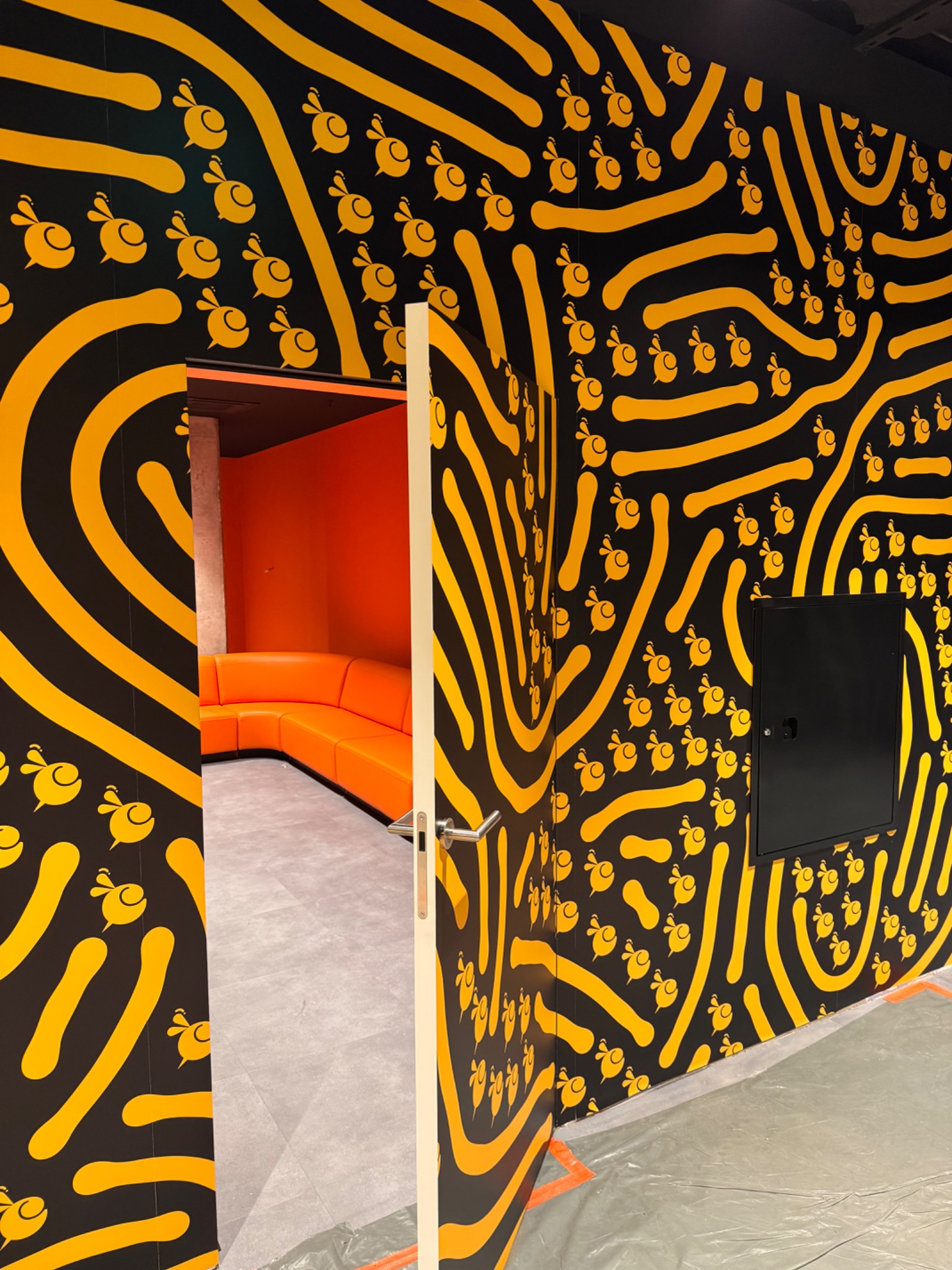 Ściana z wzorem w pszczoły i abstrakcyjne linie w kolorze żółtym na czarnym tle. Widoczne otwarte drzwi do pomarańczowego pokoju z narożną sofą.