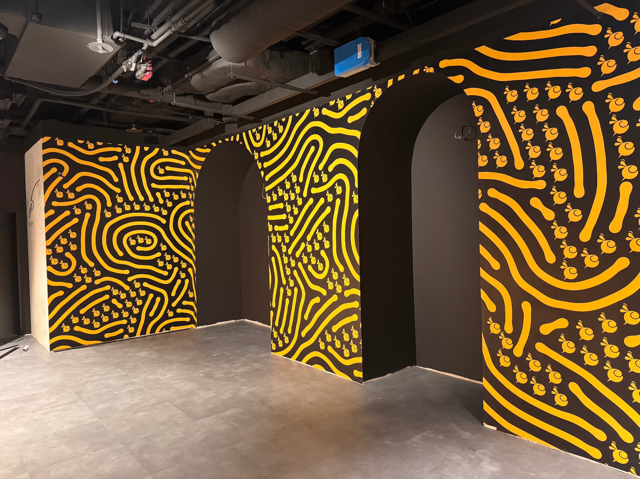 Ściana z wzorem żółtych linii i pszczół na czarnym tle, tworząca unikalny design wnętrza w Sulejówku. Widoczne łuki i instalacje pod sufitem.