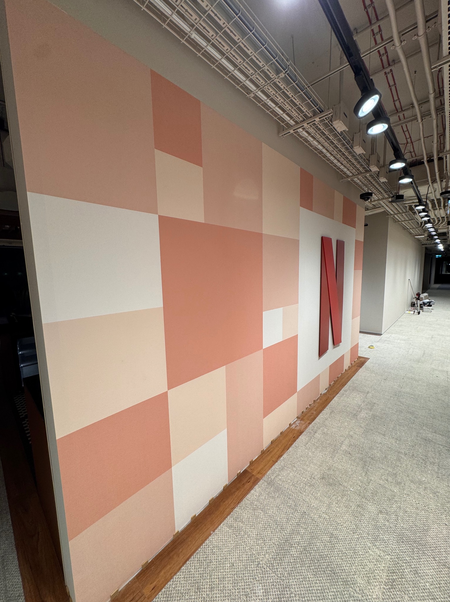 Ściana z geometrycznym wzorem w odcieniach różu i beżu, z czerwonym logo 'N'. Widok na korytarz biurowy z widoczną instalacją pod sufitem.