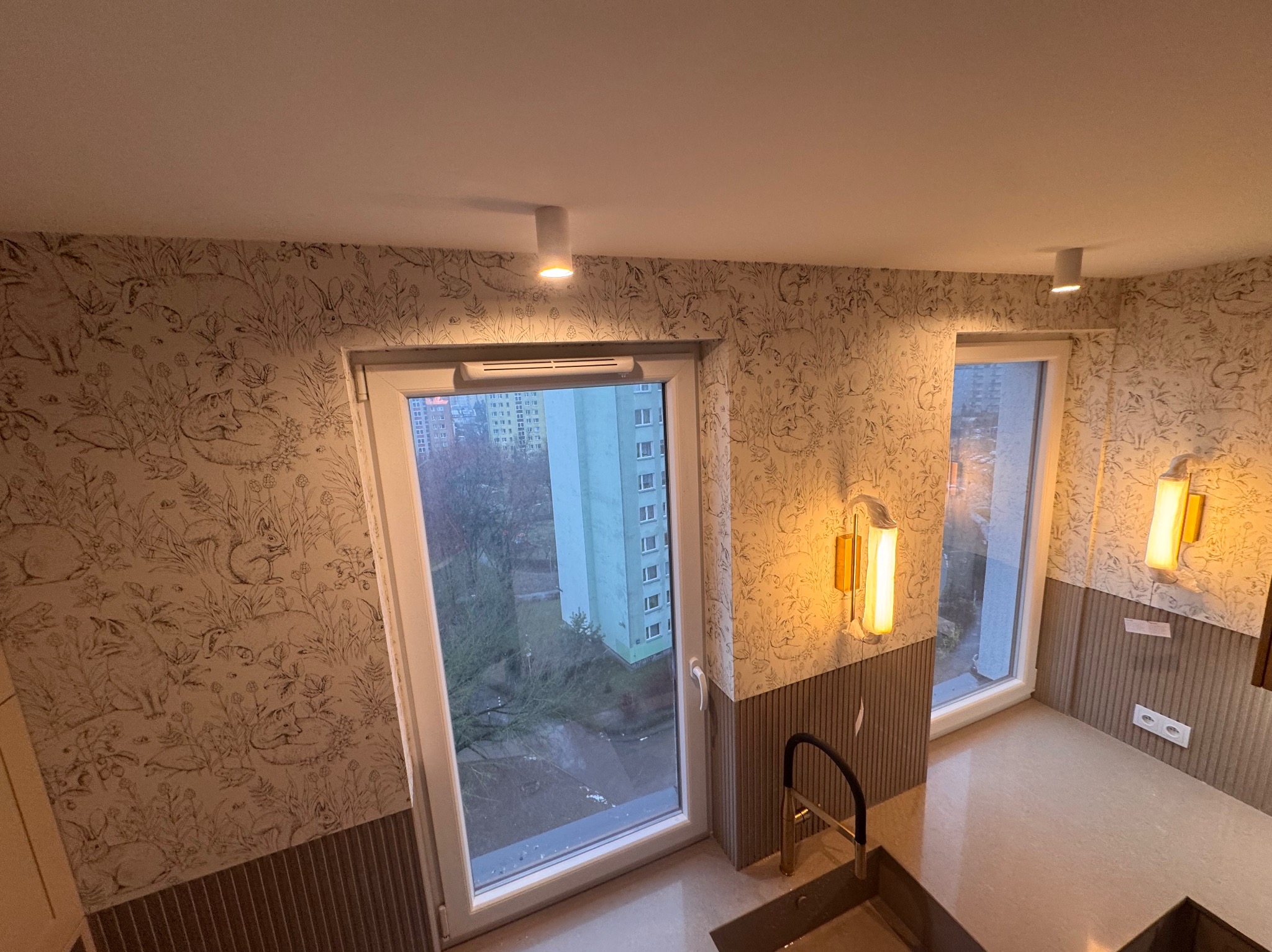 Kuchnia po remoncie: ściany zdobi tapeta w leśne motywy, okno z widokiem na blok, nowoczesne oświetlenie i armatura. Wykończenie z dbałością o detale.