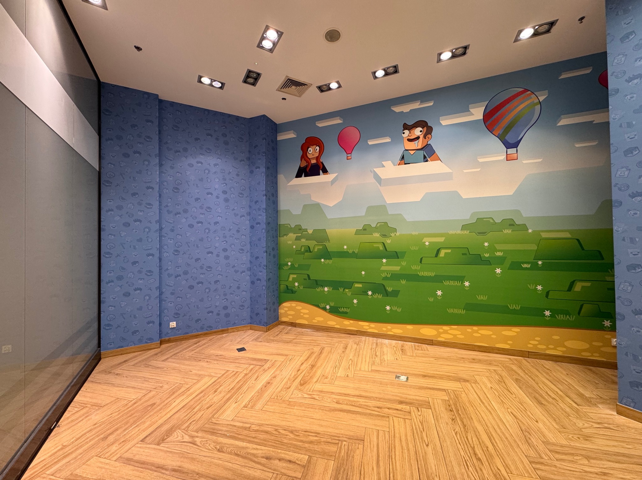 Pokój z tapetą w dziecięcym stylu: postacie z kreskówek i balony na tle błękitnego nieba i zielonych wzgórz. Podłoga z parkietu ułożonego w jodełkę.