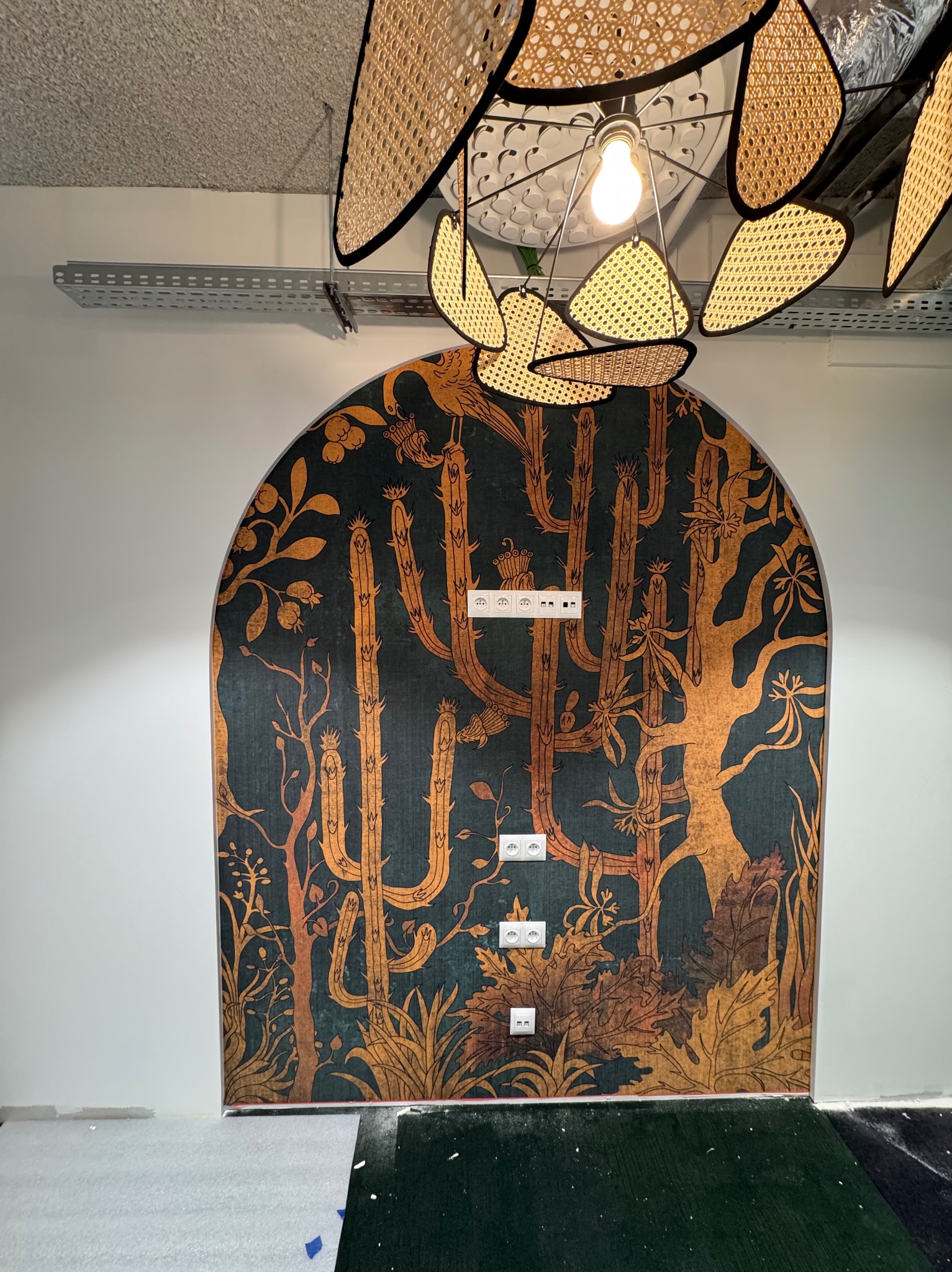 Tapeta w łuk z motywem kaktusów i roślin w odcieniach zieleni i brązu, z widocznymi gniazdkami elektrycznymi, oświetlona dekoracyjną lampą z rattanu.