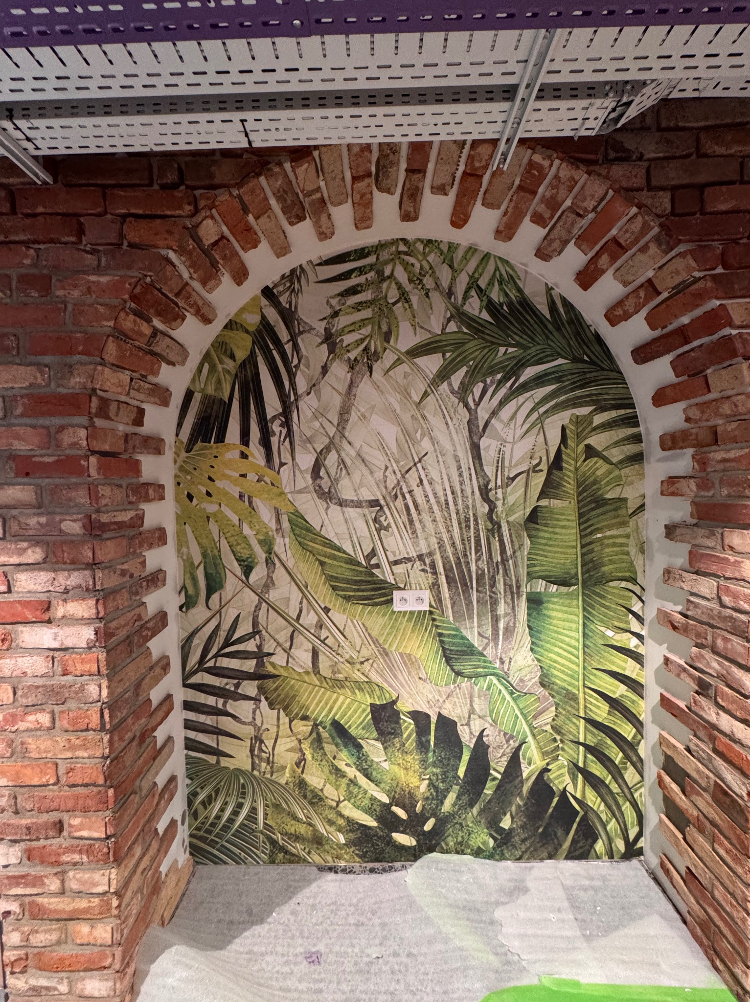 Aranżacja wnętrza: ceglany łuk z tropikalną tapetą w tle. Widoczne gniazdka elektryczne na tapecie i fragment sufitu z instalacją. Nietypowe połączenie materiałów.