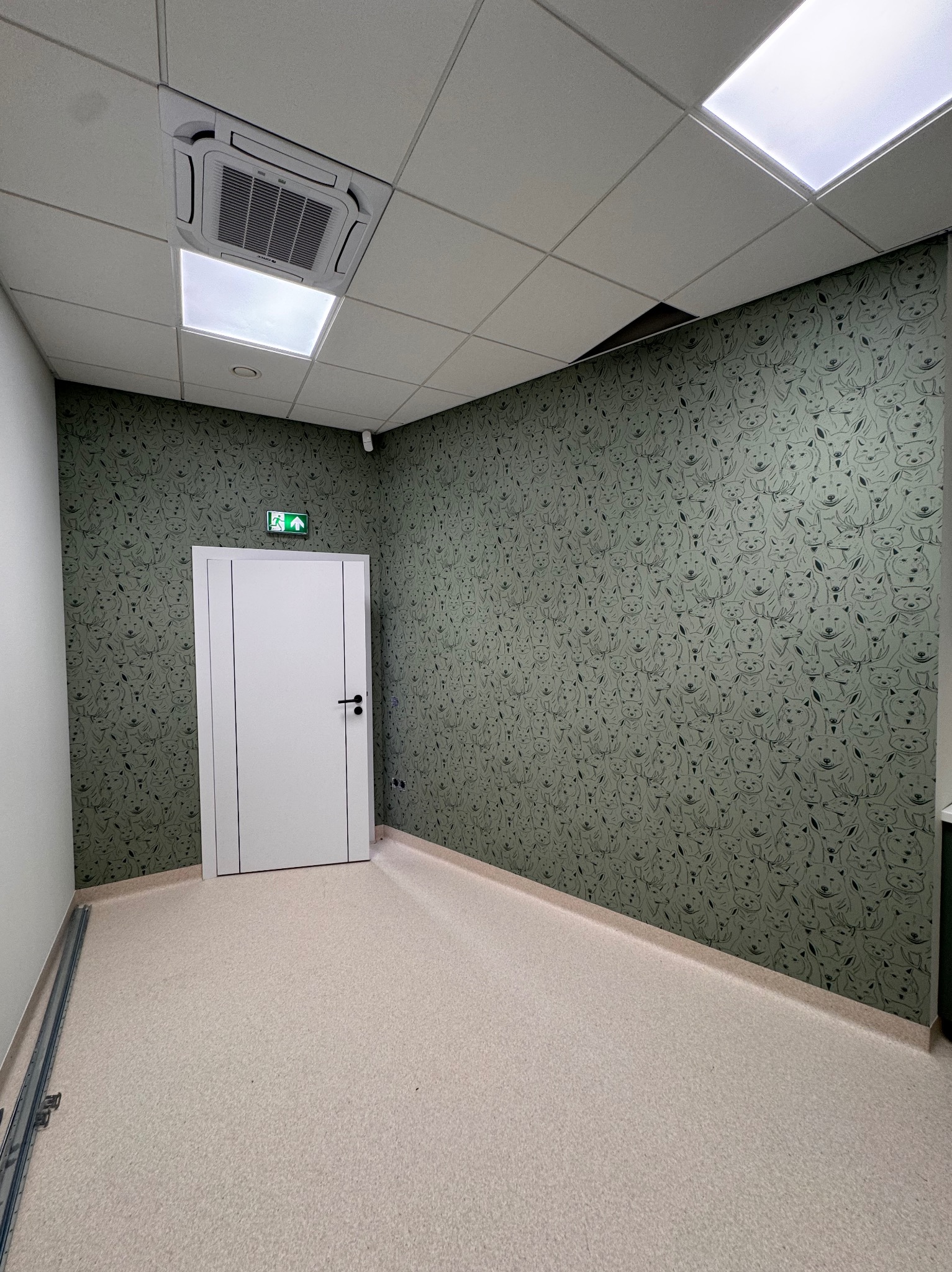 Wnętrze z tapetą w motywie zwierzęcym, białe drzwi z zielonym znakiem ewakuacyjnym, jasna podłoga i sufit kasetonowy z klimatyzacją. Nietypowy wzór tapety.