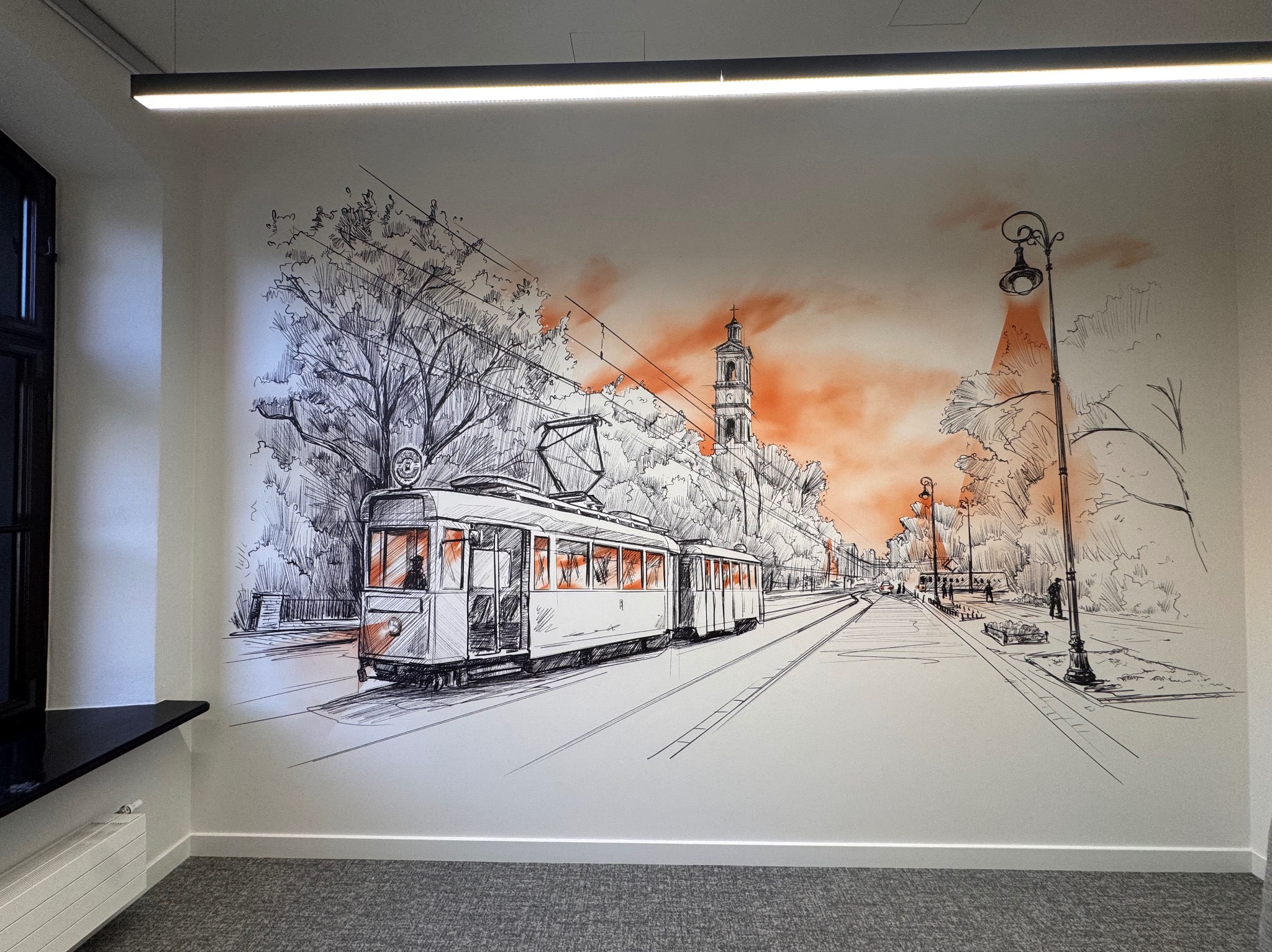 Ściana z ręcznie rysowanym muralem przedstawiającym tramwaj na tle miejskiego krajobrazu z elementami pomarańczowego koloru. Widoczne detale architektoniczne i drzewa.