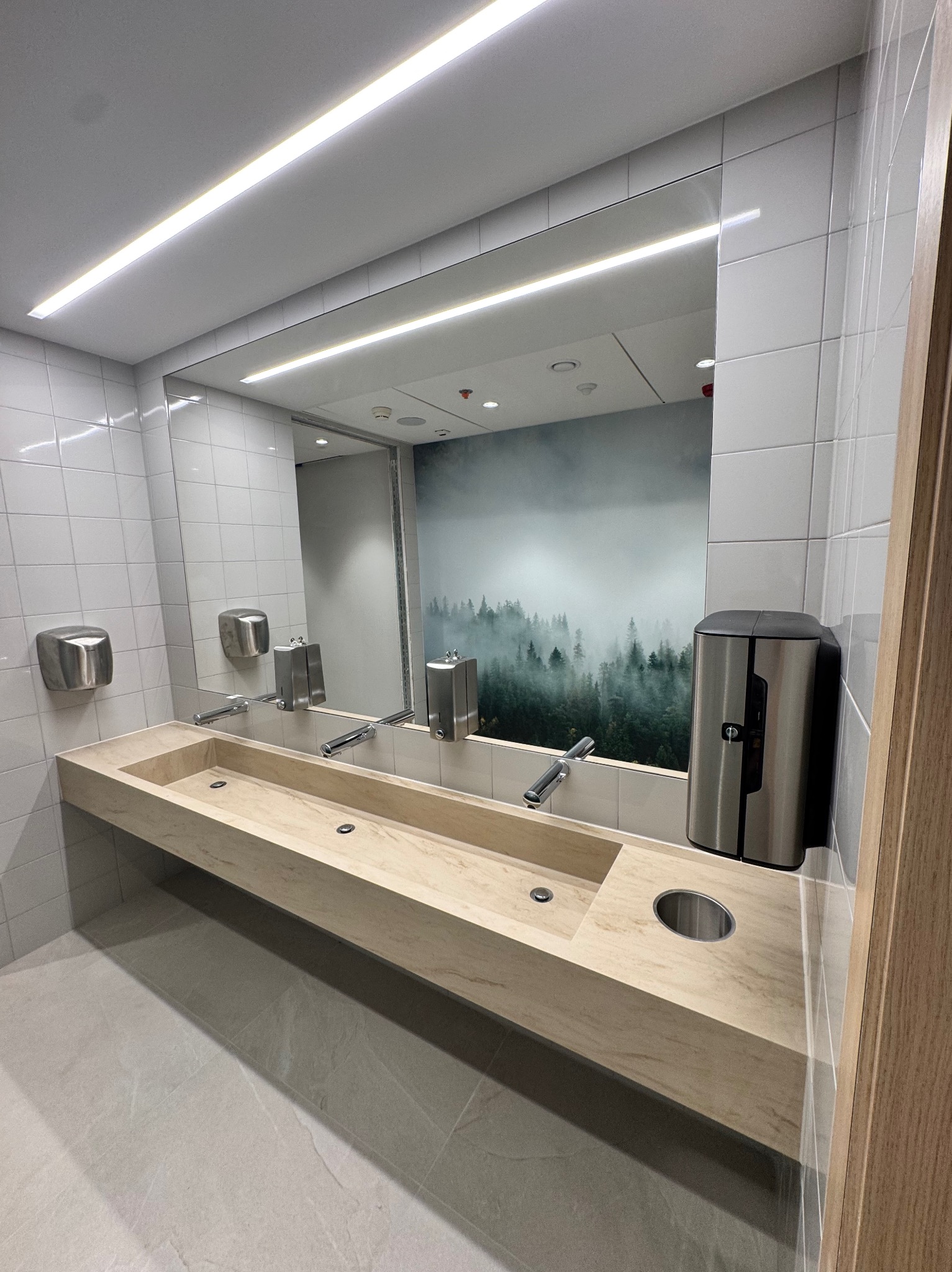 Nowoczesna łazienka publiczna z długą, kamienną umywalką, srebrnymi dozownikami i lustrem odbijającym tapetę z motywem lasu we mgle. Minimalistyczny design.