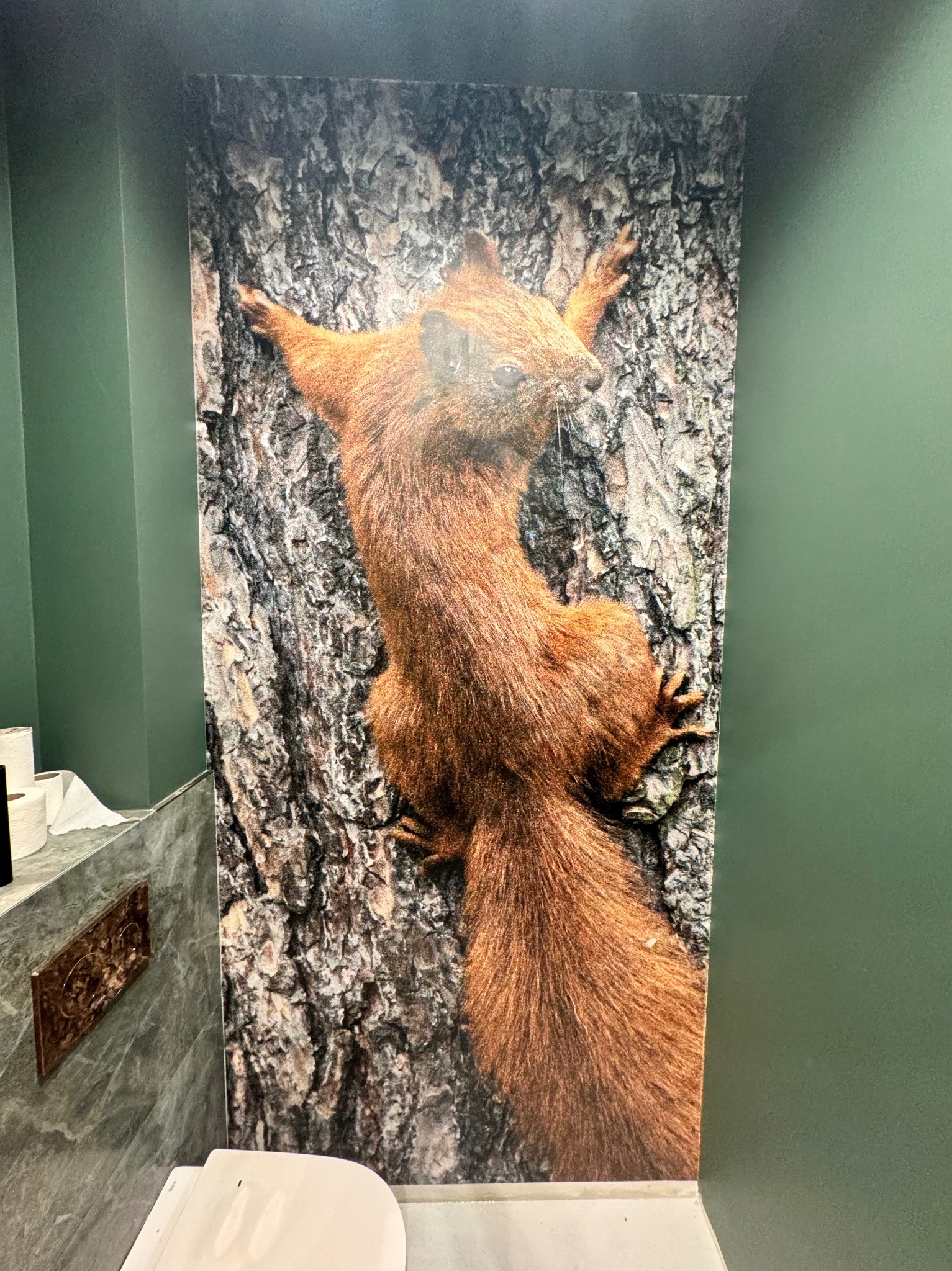 Fototapeta z wiewiórką na ścianie w łazience, obok toaleta i marmurowe wykończenie. Zielone ściany i nietypowy wzór tapety tworzą unikalny wystrój.