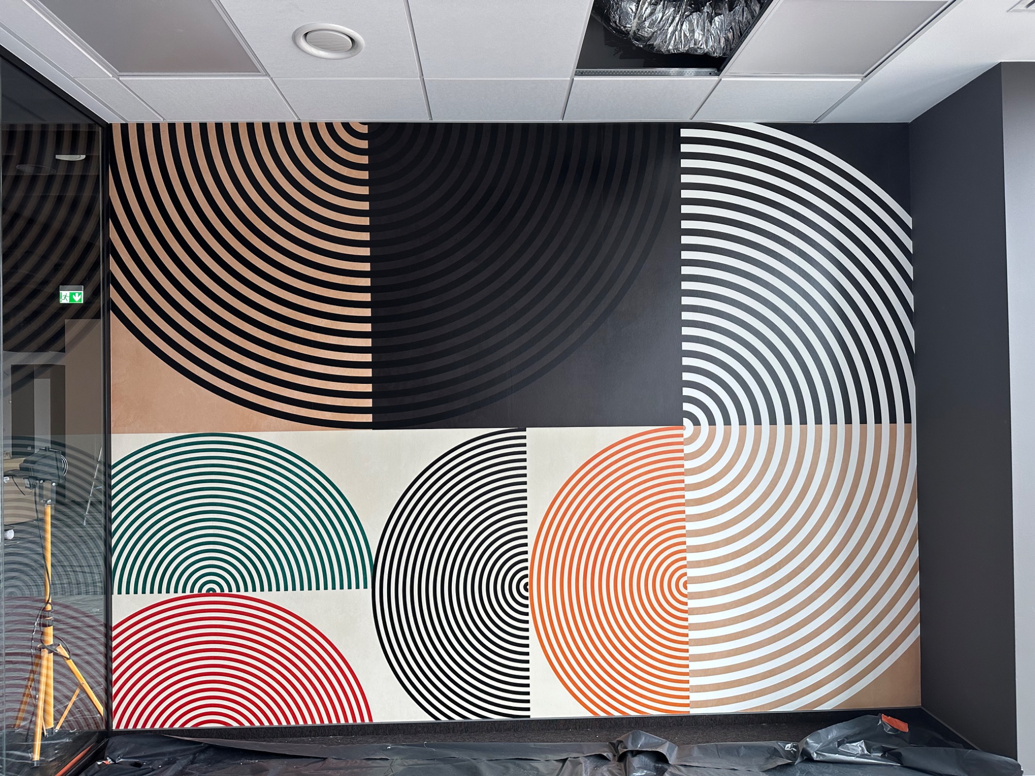 Ściana biurowa z geometrycznym wzorem z półkolistych pasów w kolorach czarnym, białym, beżowym, zielonym i czerwonym, widoczny reflektor na statywie i folia ochronna na podłodze.
