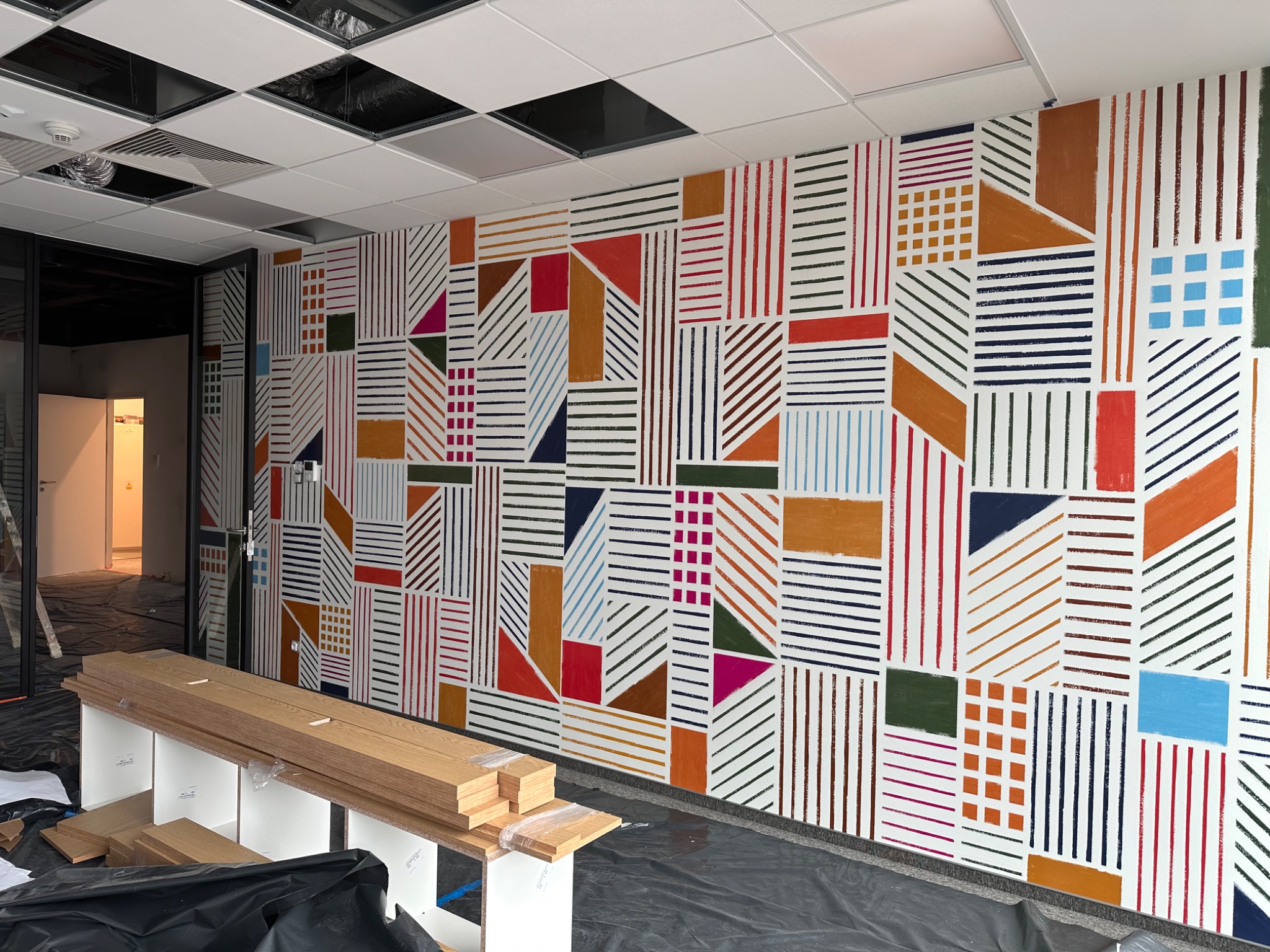 Ściana z geometrycznym, ręcznie malowanym wzorem w różnych kolorach, widoczne przygotowane deski podłogowe, folia ochronna na podłodze, sufit kasetonowy.