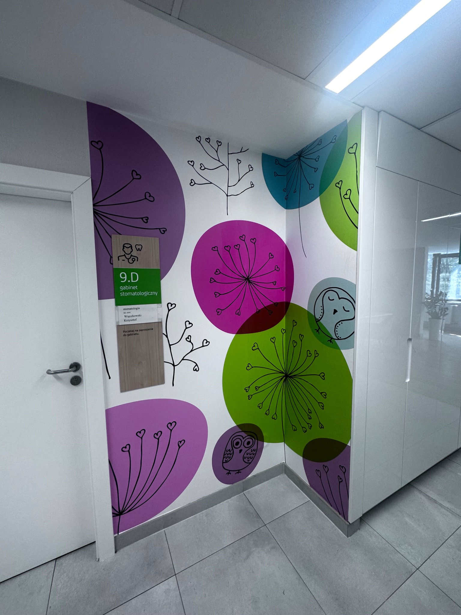 Ściana w gabinecie stomatologicznym z abstrakcyjnymi, kolorowymi wzorami roślinnymi i motywami zwierzęcymi, takimi jak sowy, oraz tabliczka informacyjna z numerem gabinetu 9.D i nazwiskiem lekarza...