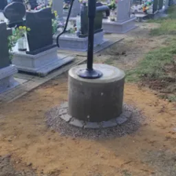 Czarna pompa ręczna na betonowej podstawie otoczonej drobnymi kamieniami na cmentarzu, w tle nagrobki i drzewa.