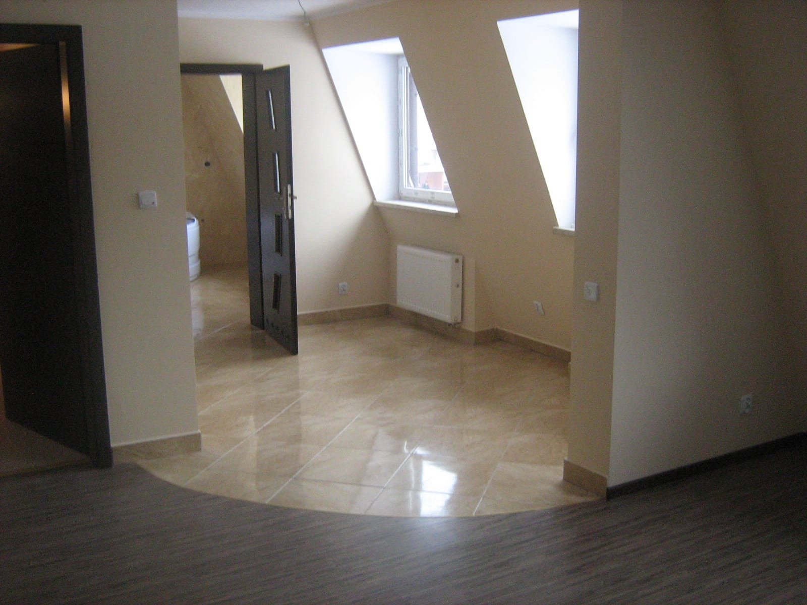 Wykończone wnętrze poddasza z beżowymi płytkami podłogowymi, grzejnikiem pod oknem dachowym i otwartymi ciemnymi drzwiami do sąsiedniego pomieszczenia z widoczną białą wanną.